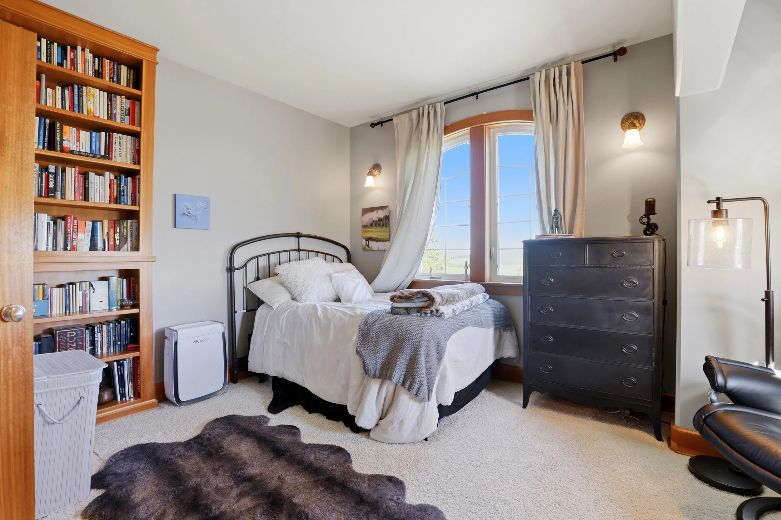 A bright bedroom with a bed next to a large window, a black dresser, and a bookshelf filled with books. There are cream-colored curtains, wall-mounted lamps, and a furry rug on the beige carpeted floor.