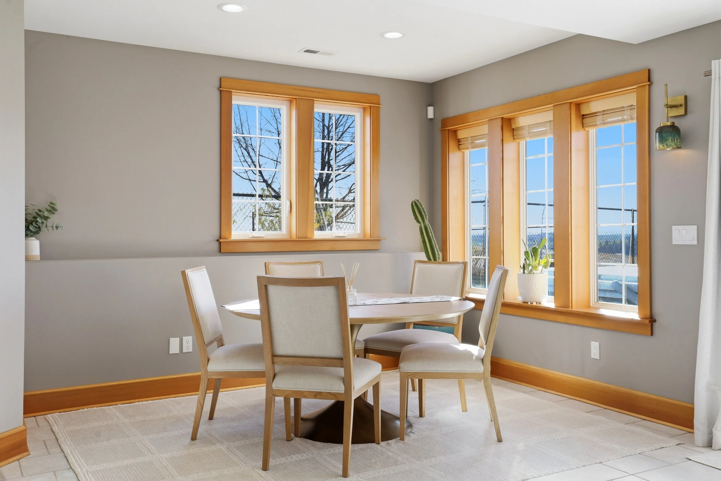 A dining area with a round wooden table and four beige chairs. Large windows with wooden trim let in natural light, and outside, a tree is visible. There are potted plants and a white curtain in the room.