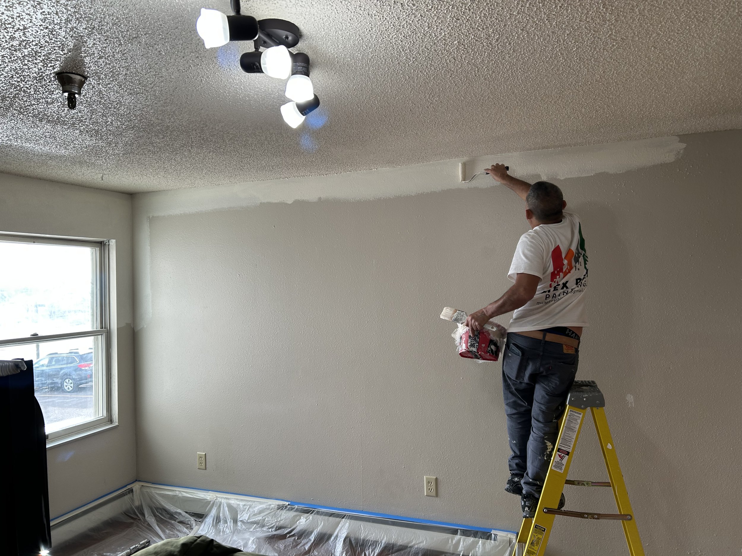 A person standing on a yellow step ladder painting the upper part of a white wall near the ceiling in a room during renovations.