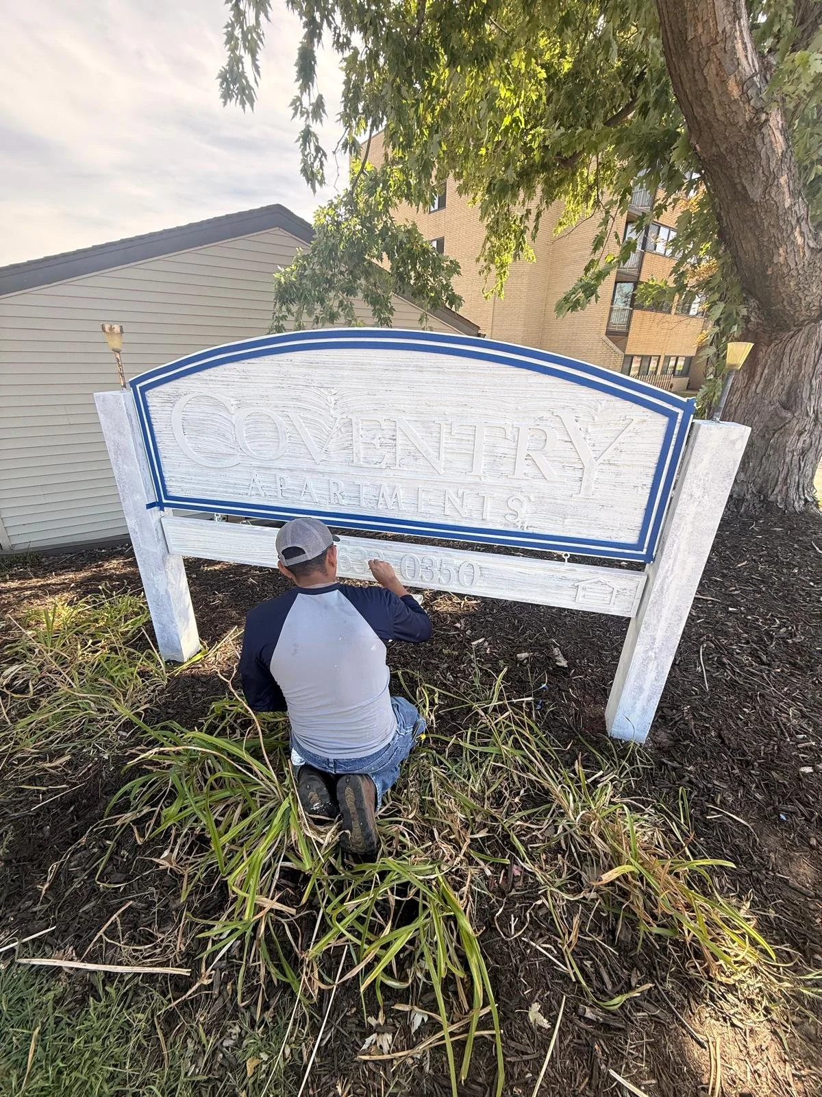 A man kneeling and painting a large white sign for a complex called "Coventry Apartments" with a phone number, surrounded by mulch and plants, with trees and buildings in the background.