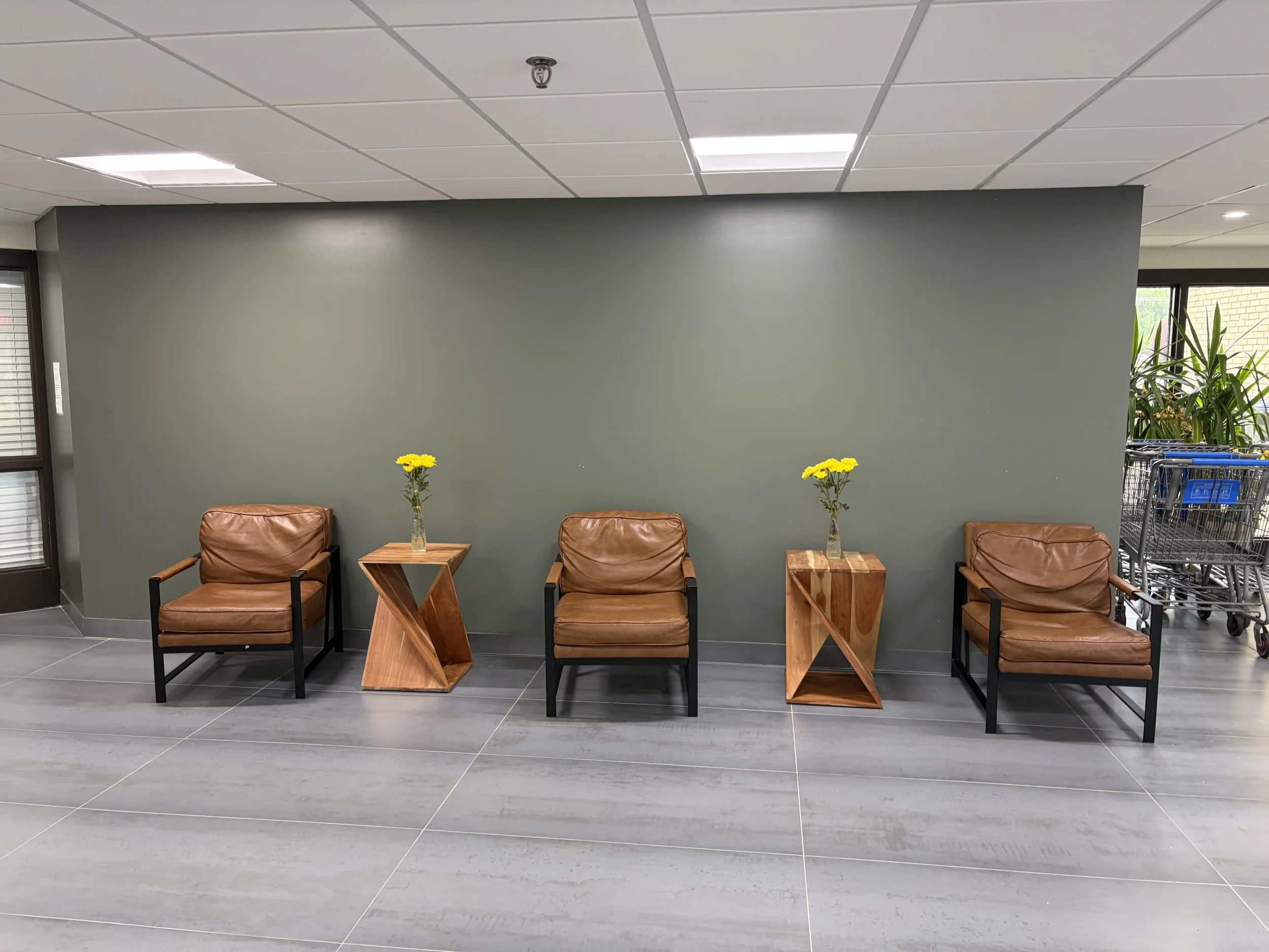 Interior of a waiting area with a light green wall, brown leather chairs, small wooden side tables with yellow flowers, and shopping carts to the right.
