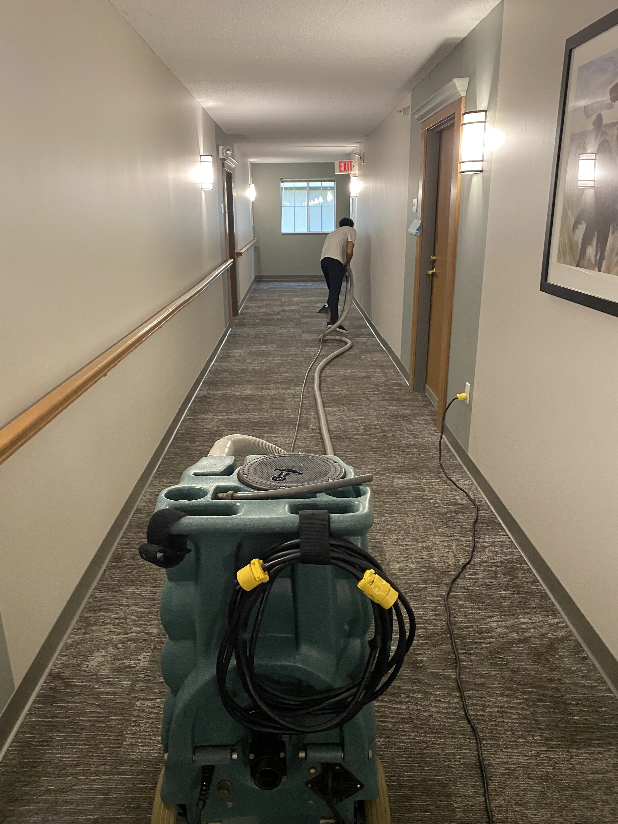 A person is vacuuming a carpeted hallway with a green commercial vacuum cleaner, viewed from behind.