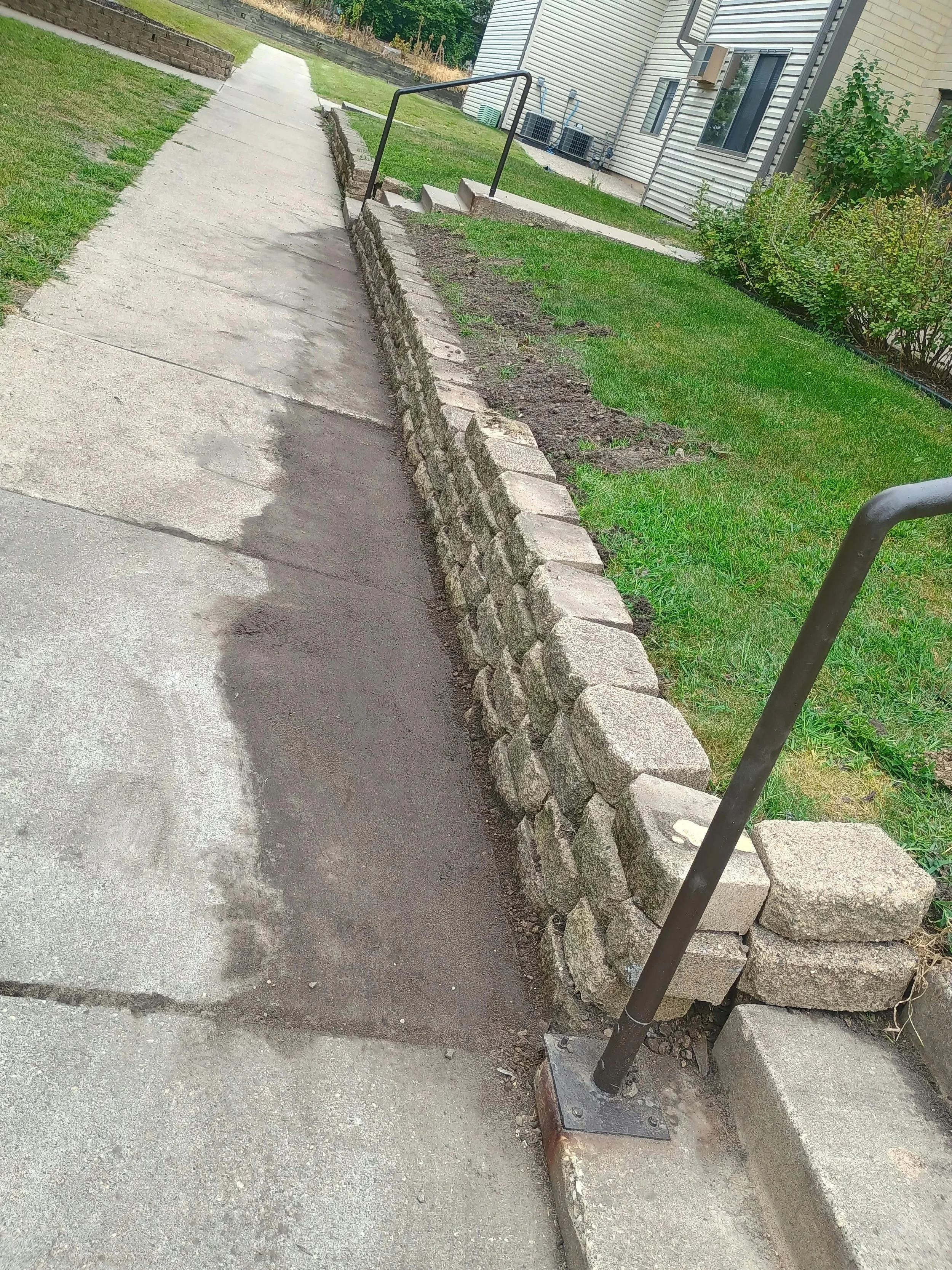 A sidewalk with a small stone retaining wall on the right, a black metal handrail, a grassy area, and a building with air conditioning units and windows in the background.