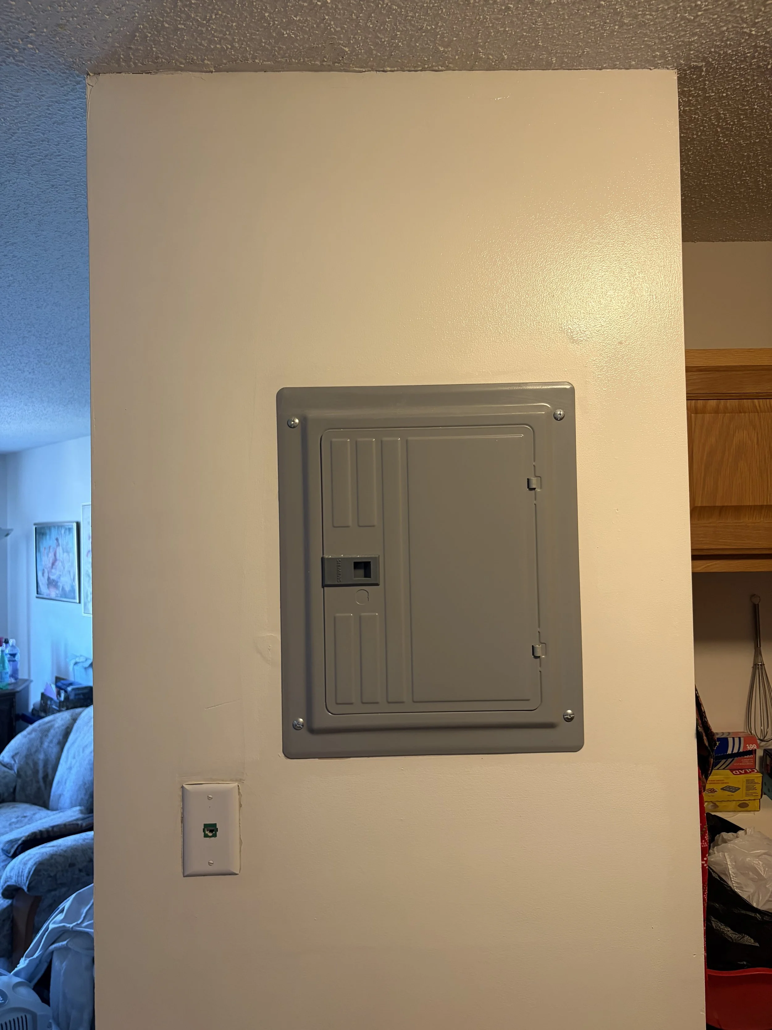 A beige wall with a gray electrical panel, a white switch plate with a green sticker, and a corner of a room with a wooden cabinet and a cluttered corner.