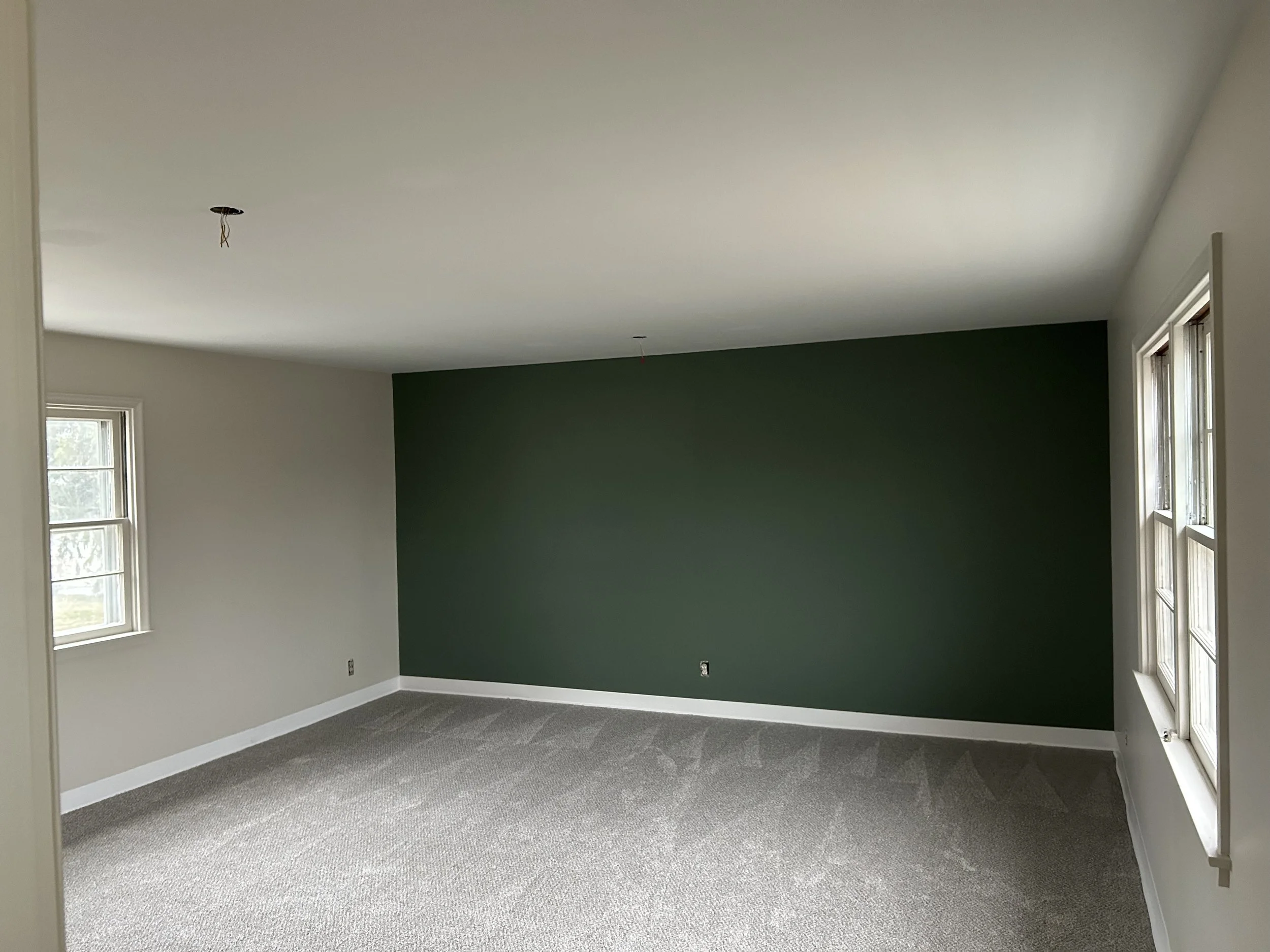An empty room with beige walls, a green accent wall, large windows, and beige carpet flooring, with electrical outlets awaiting installation.