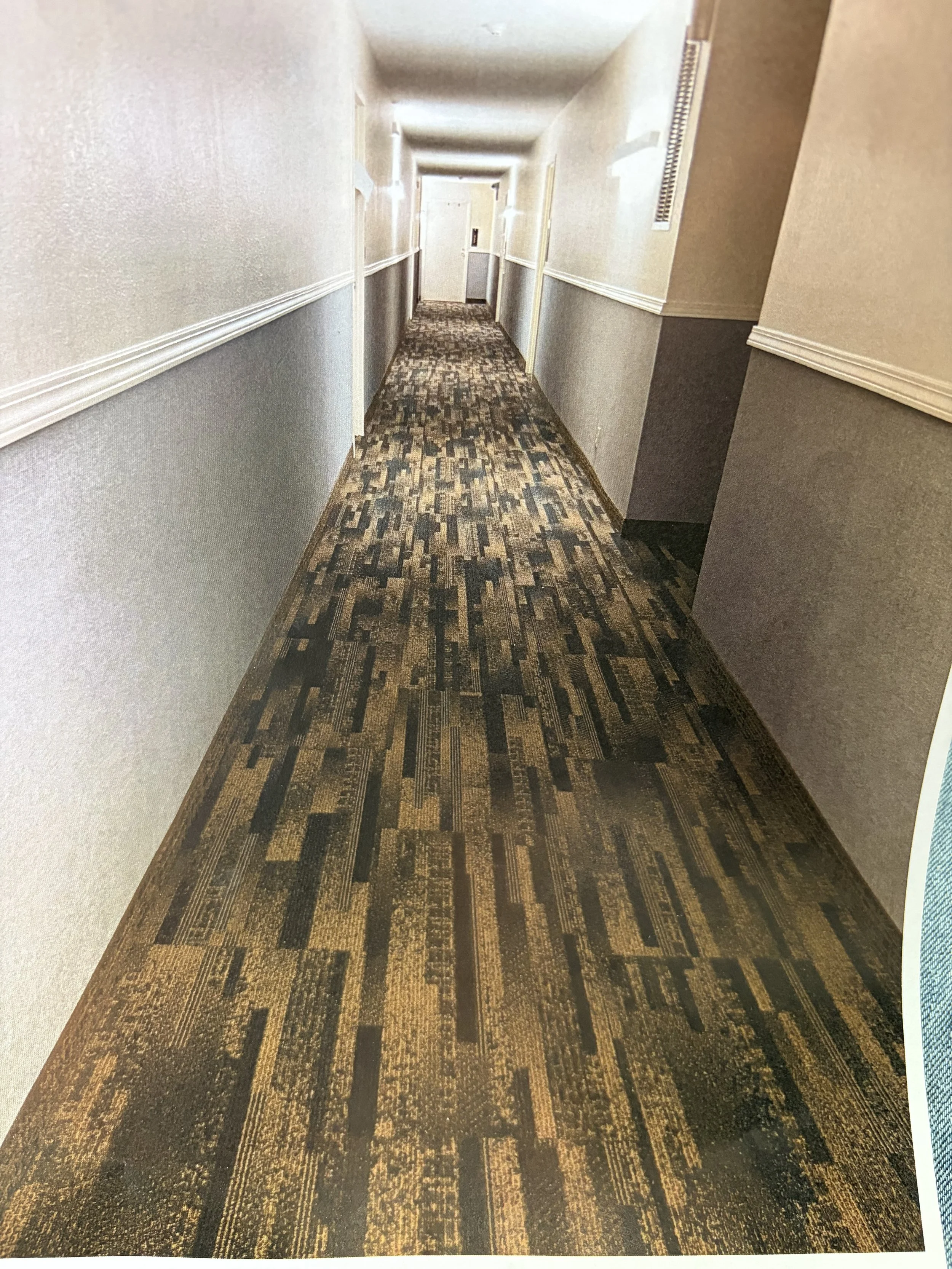 Long hotel hallway with patterned carpet, beige and light gray walls, white baseboards, and ceiling; several closed doors along both sides.