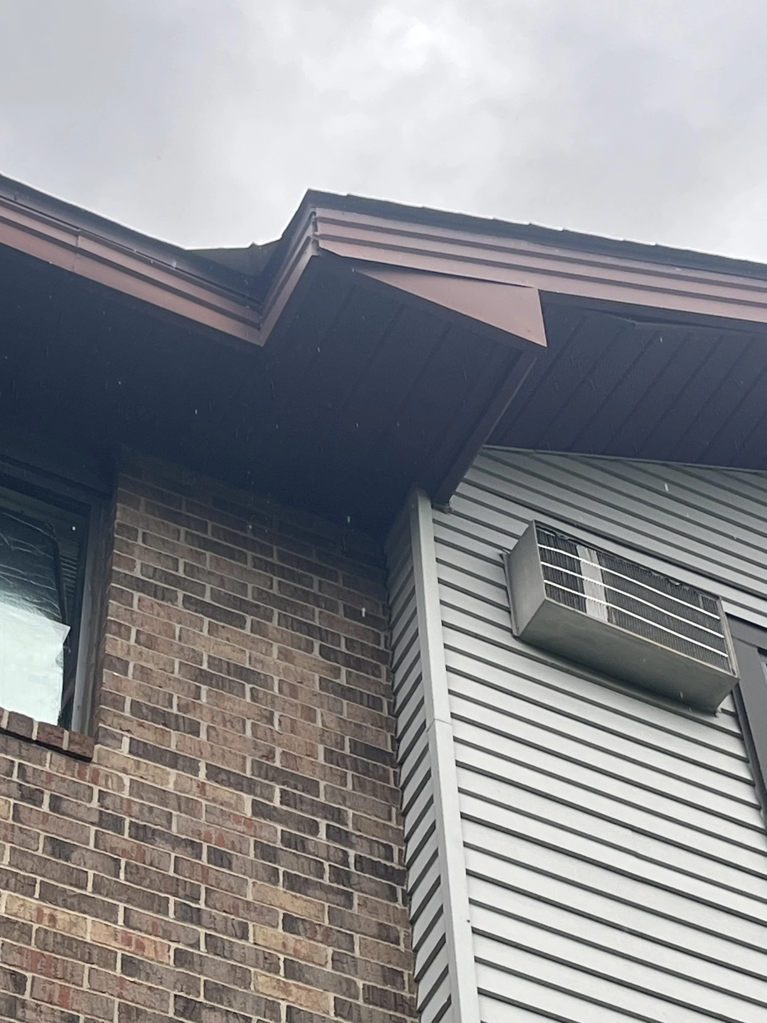 Close-up of a building corner showing brick wall, vinyl siding, and a roof with metal trims and vents, under a cloudy sky.