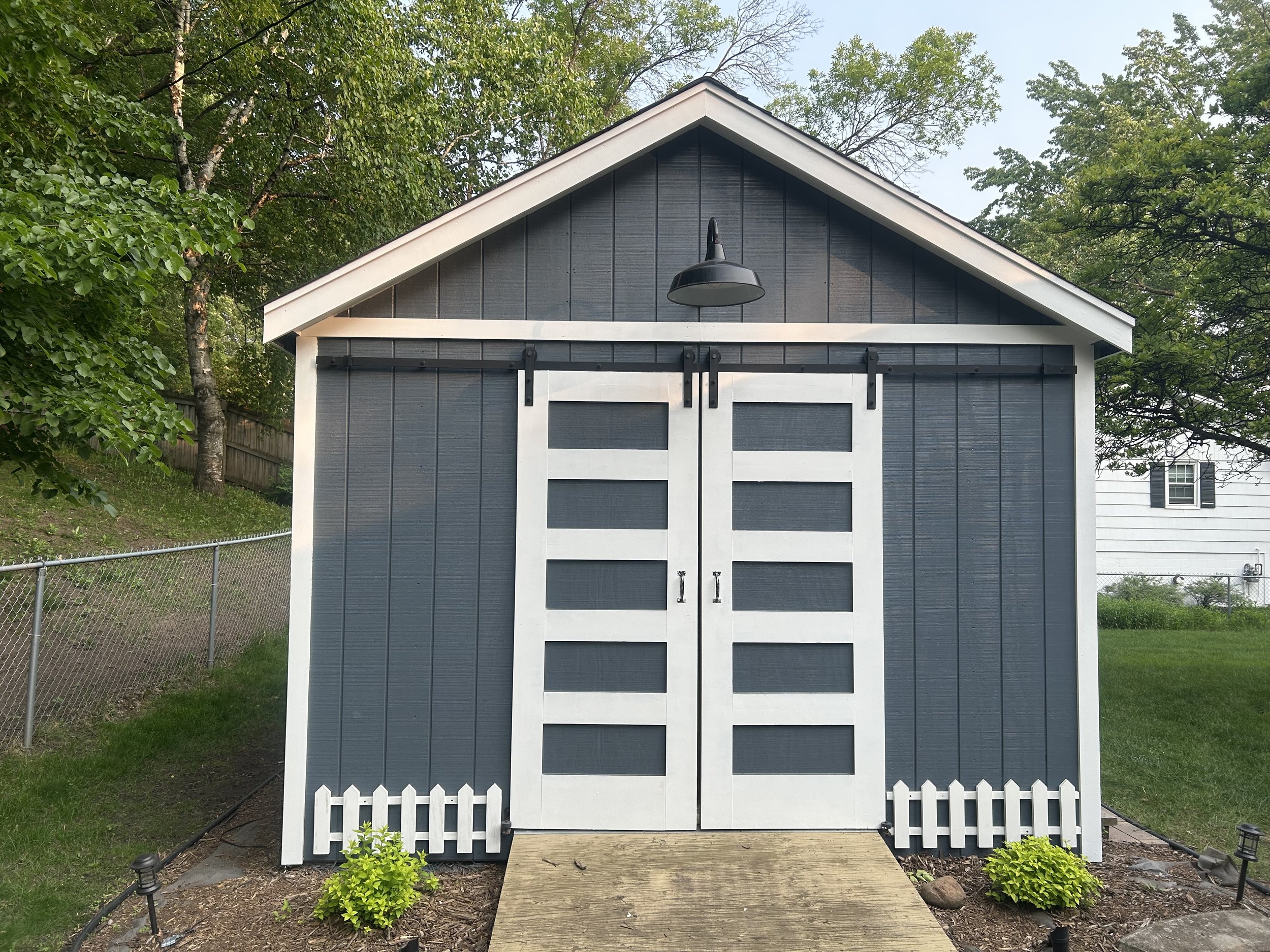 A small backyard shed with a blue wooden exterior, white trim, and black sliding barn doors. The shed has a gabled roof with white edges, a black outdoor light fixture, a small wooden step, and is surrounded by a manicured garden with small bushes, a white picket fence, and a chain-link fence. Trees and a neighboring house are visible in the background.