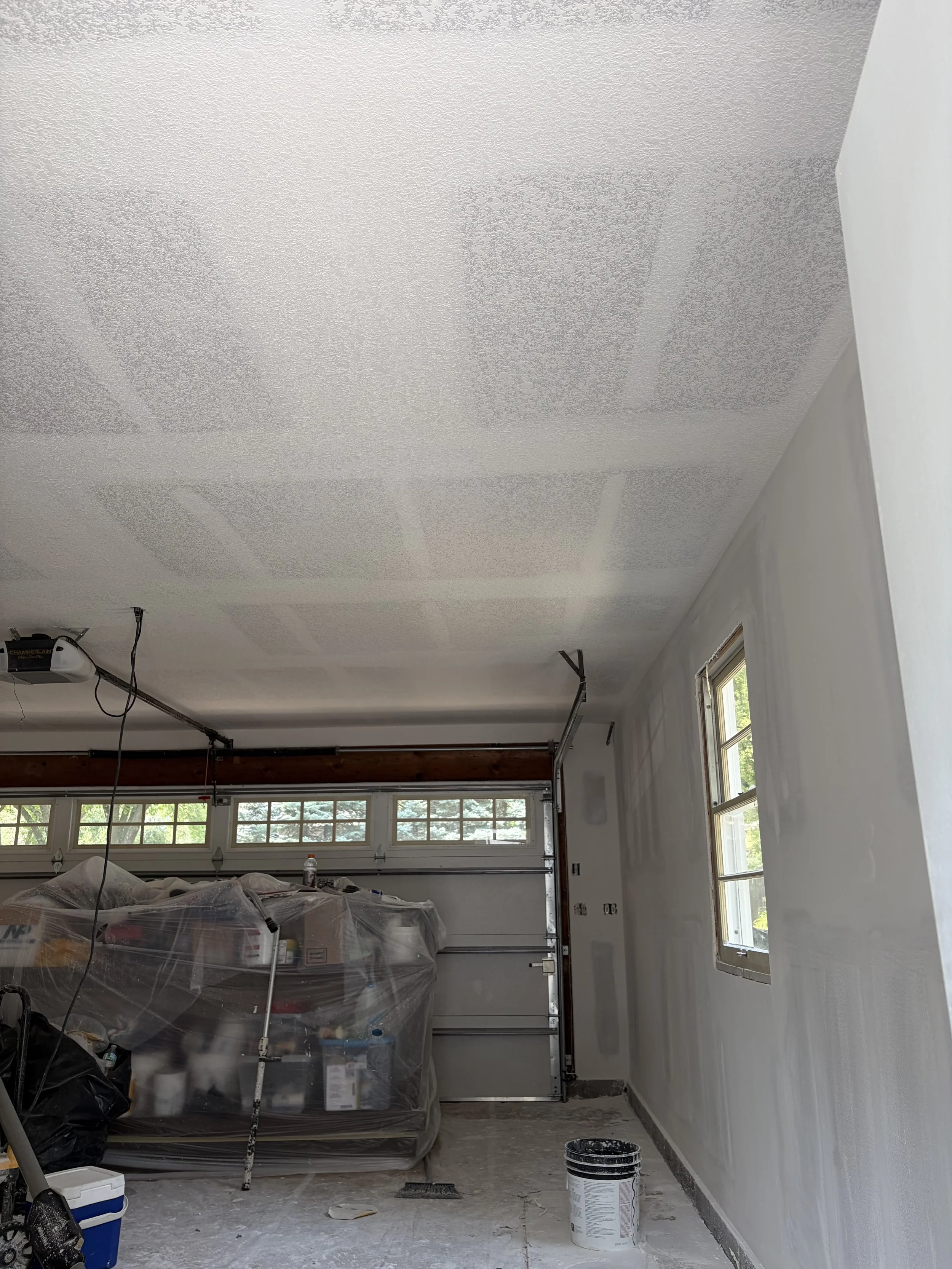 A room under renovation with a textured white ceiling and a partially painted white wall, windows along the side, plastic-covered furniture and tools scattered on the floor, and a bucket of paint in front of the wall.