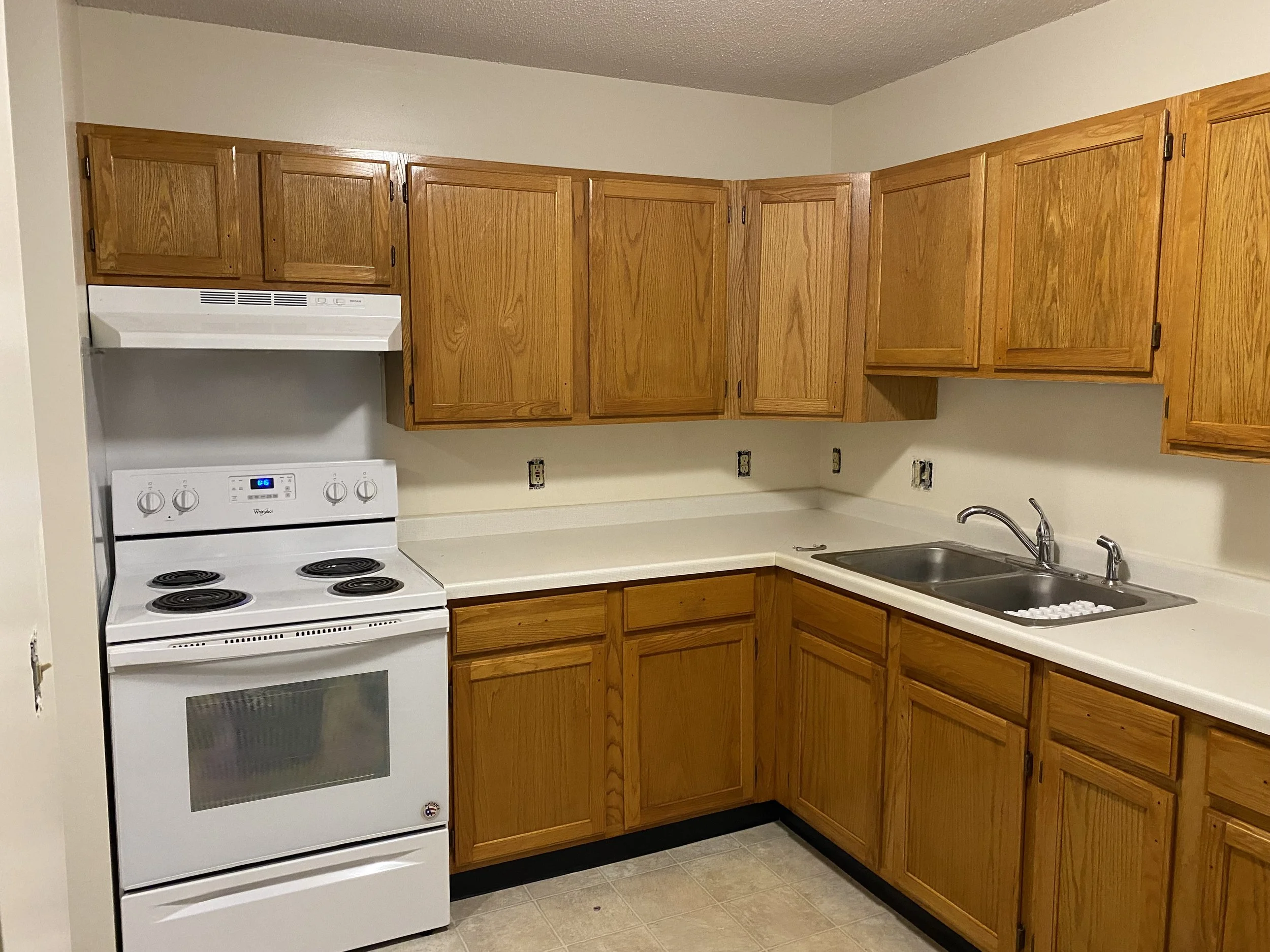 A kitchen with wooden cabinets, a white electric stove, double sink with a chrome faucet, and a beige countertop. The wall has several empty electrical outlets.