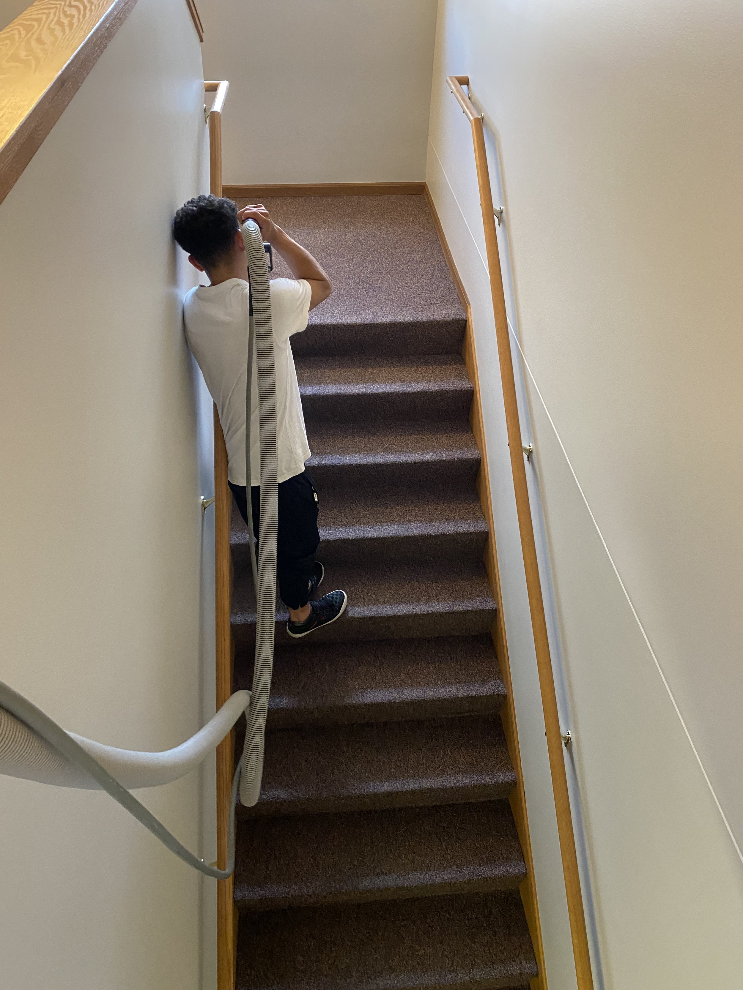 A man with curly hair wearing a white T-shirt and black pants is vacuuming brown carpeted stairs in a house. The vacuum hose is attached over his shoulder, and he is standing at the bottom of the staircase.
