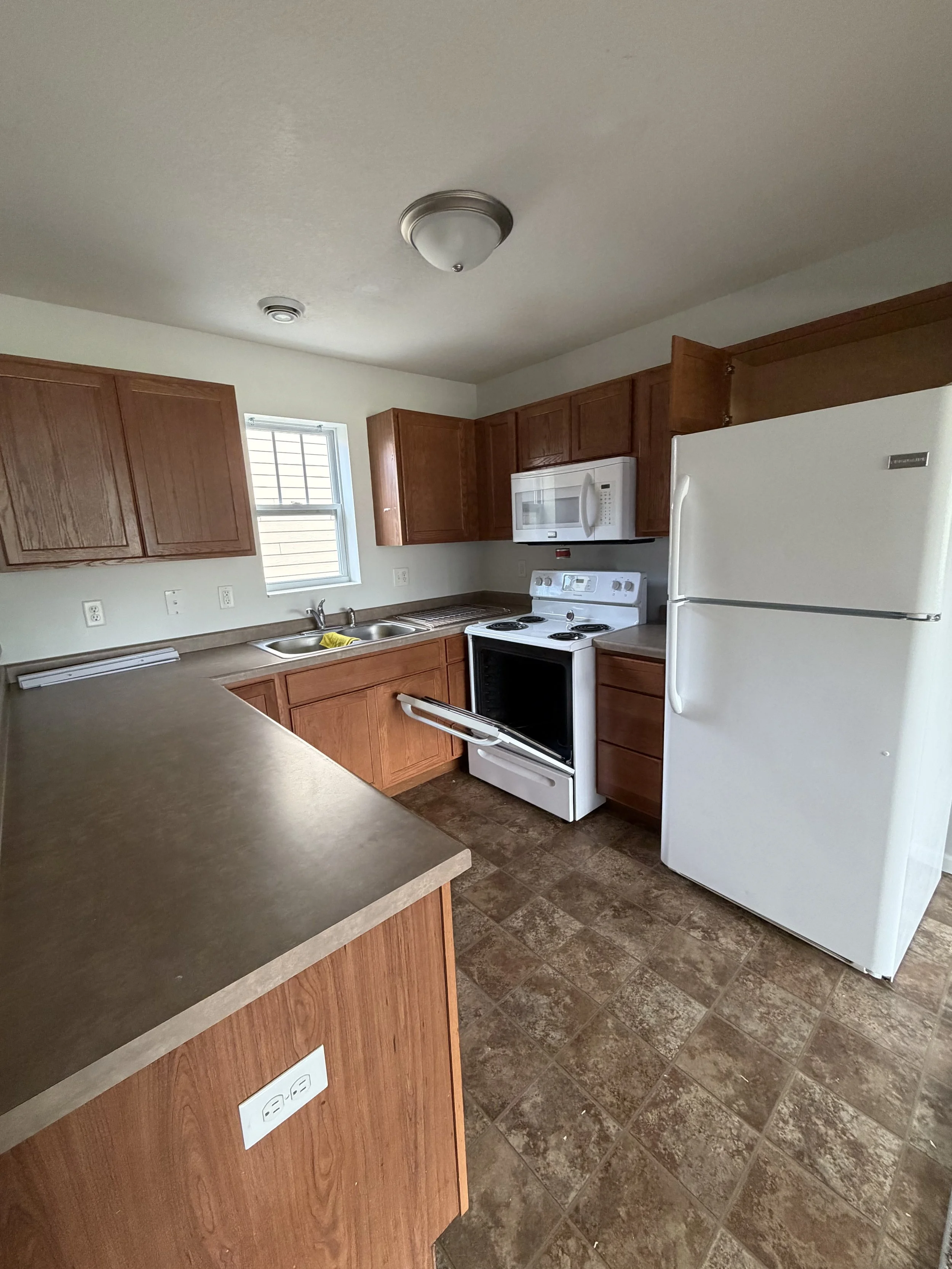 A kitchen with wooden cabinets, a white refrigerator, a white stove, a microwave, a sink under a window, and tiled flooring.
