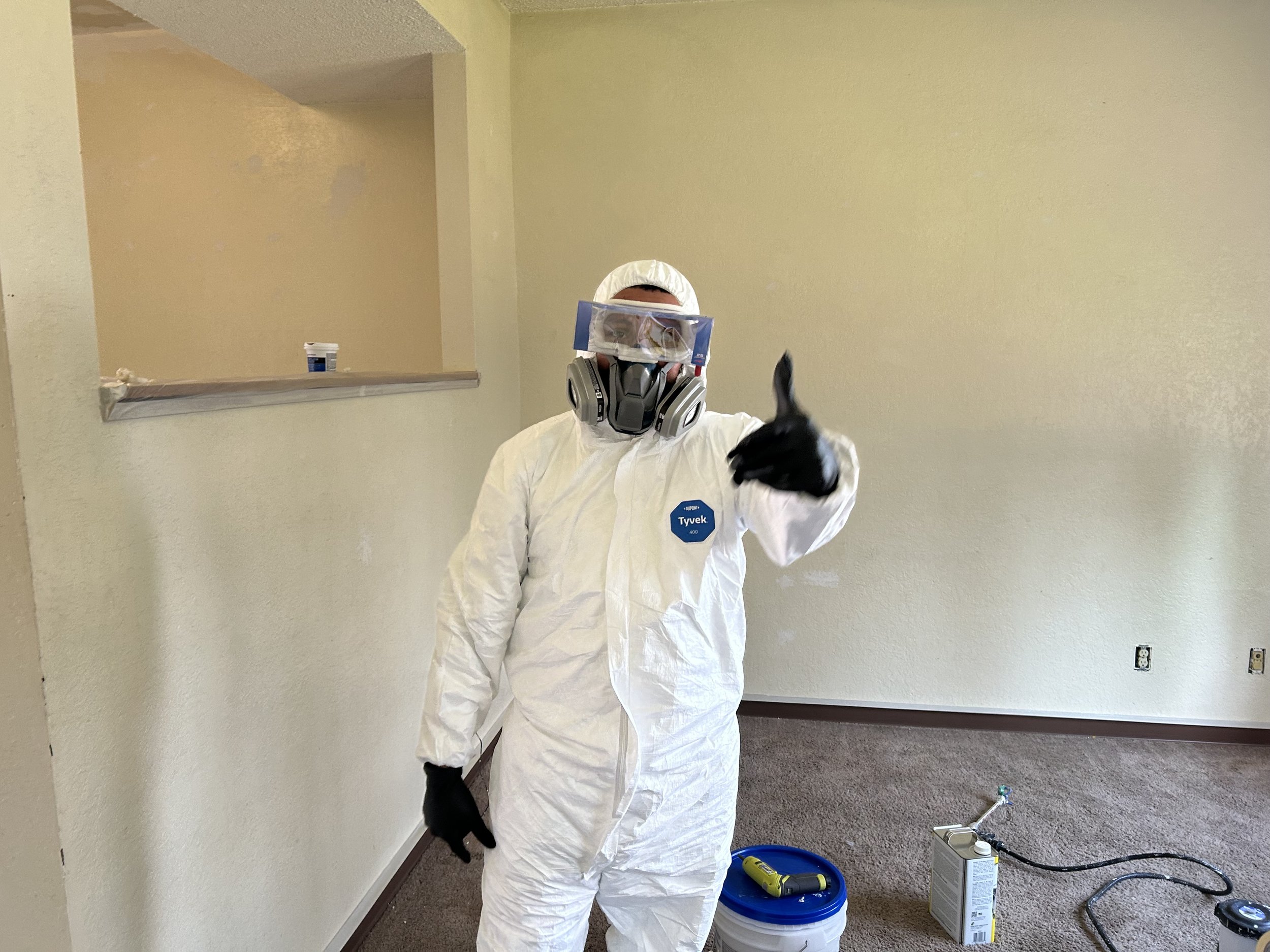 Person dressed in a white protective suit, wearing a respirator mask, gloves, and face shield, giving a thumbs up inside a room with beige walls. There are containers and tools on the floor, suggesting a cleaning or renovation activity.