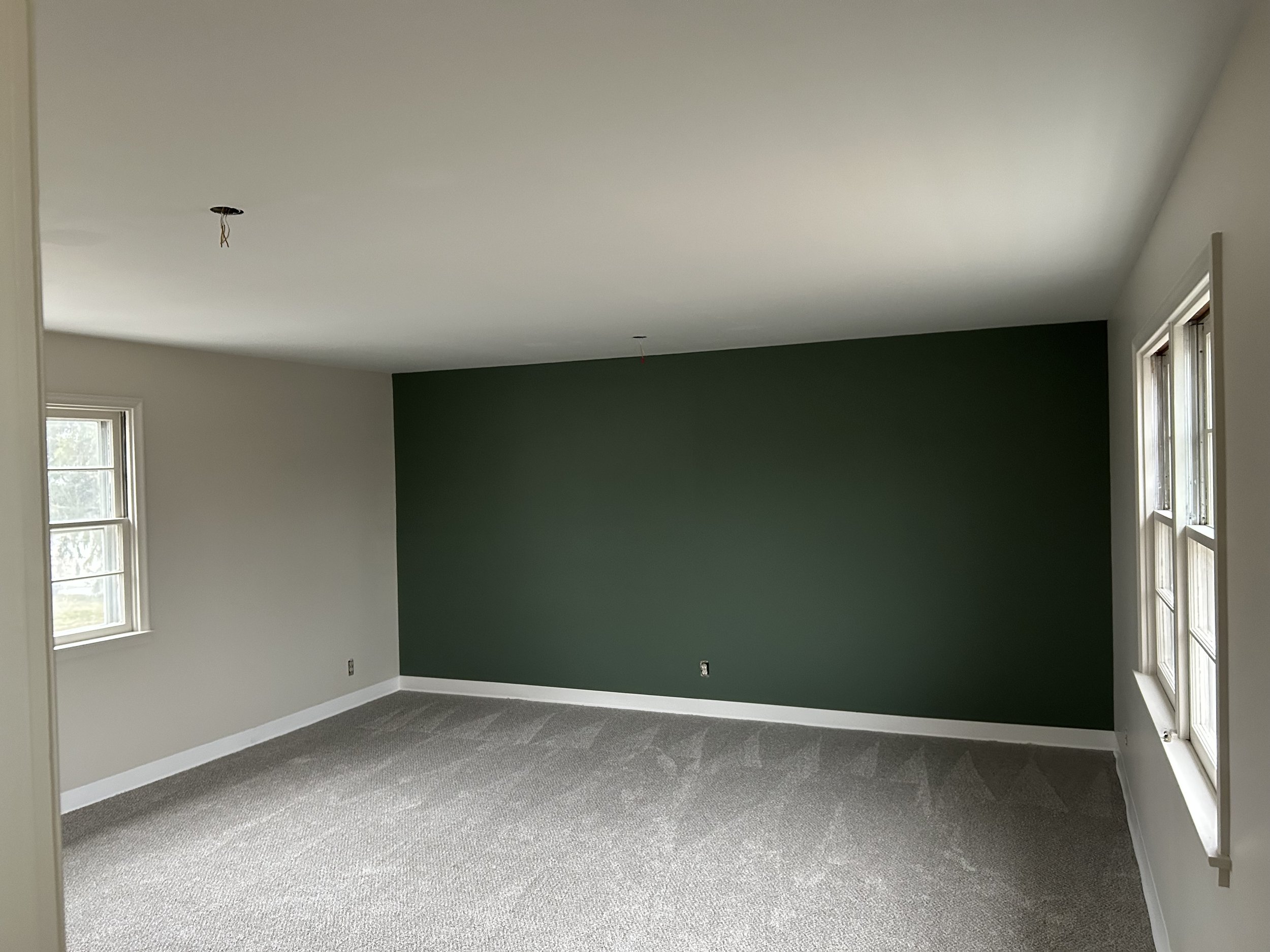 An empty room with a green accent wall, two windows, and neutral-colored carpeting.