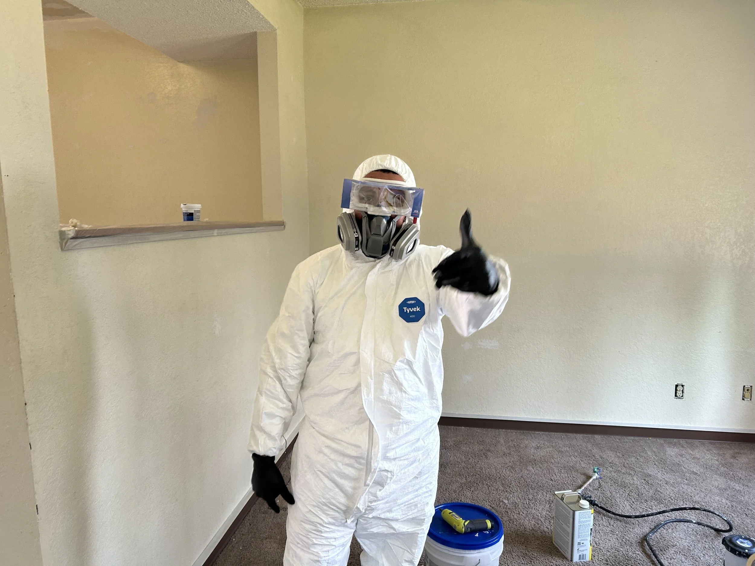 Person wearing white coveralls, gloves, a face shield, and a respirator mask giving a thumbs-up in an empty room with beige walls and gray carpet. There are painting supplies and a can of paint on the floor.