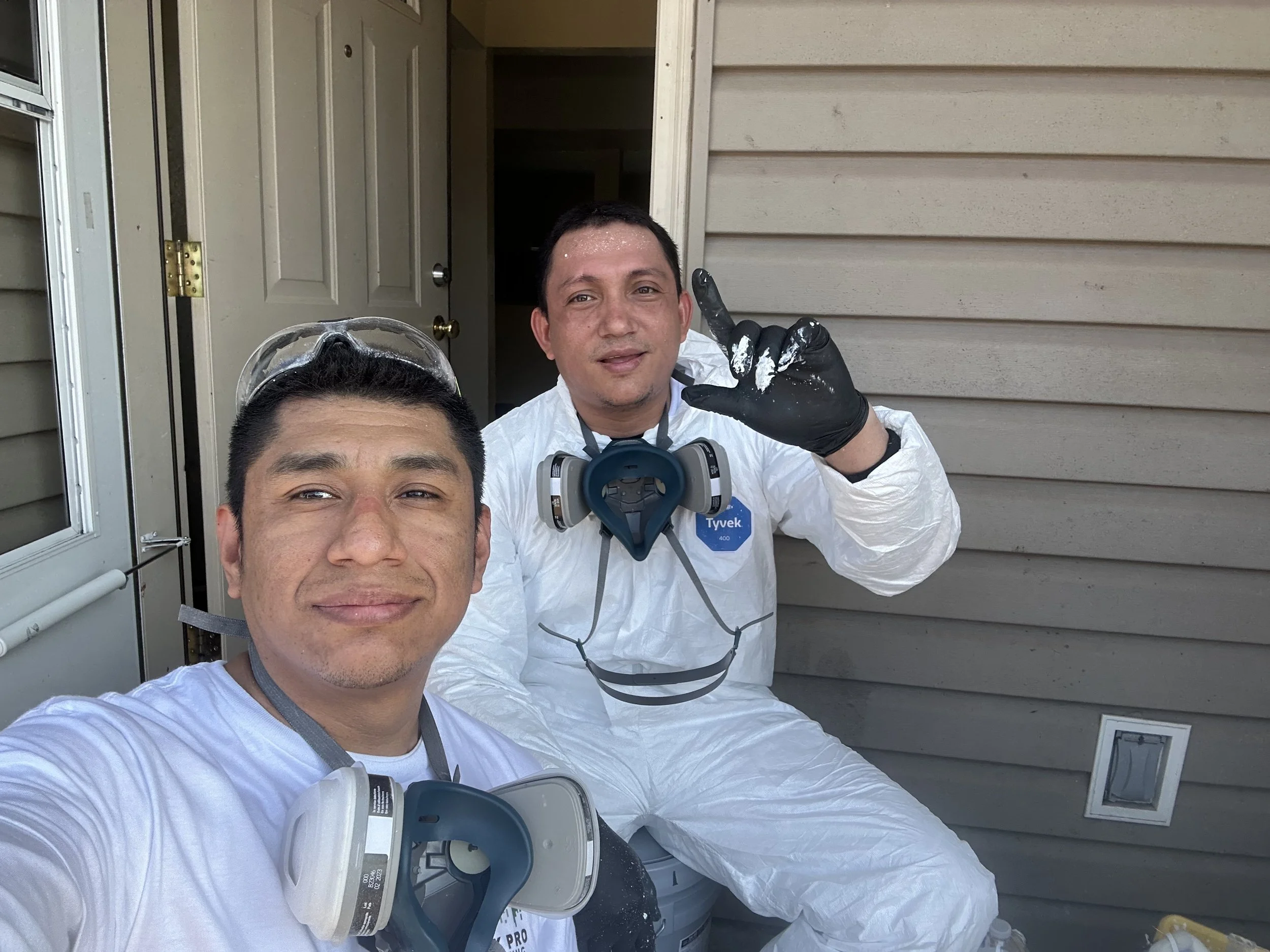 Two men wearing protective gear, including masks and gloves, taking a selfie on a porch outside a house. One man is in the foreground, and the other making a peace sign with paint on his glove.