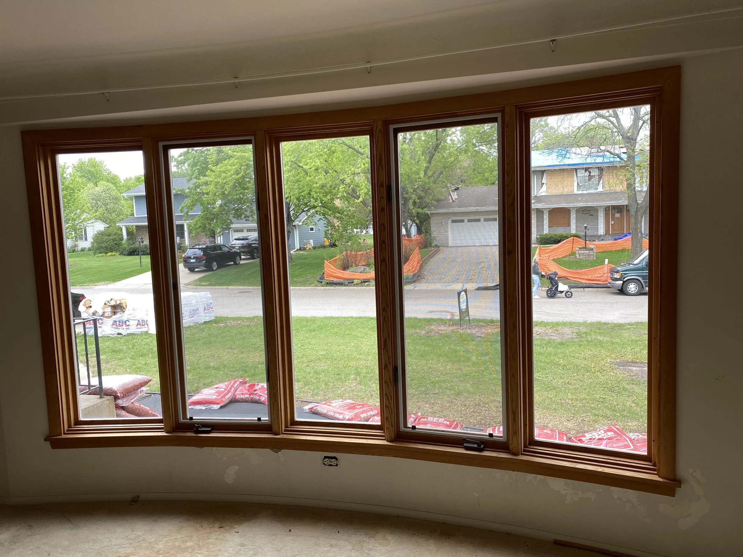 View from inside a house looking out of a new or renovated bay window with multiple glass panes, showing a suburban neighborhood with trees, houses, parked cars, and a small sidewalk, during daytime.