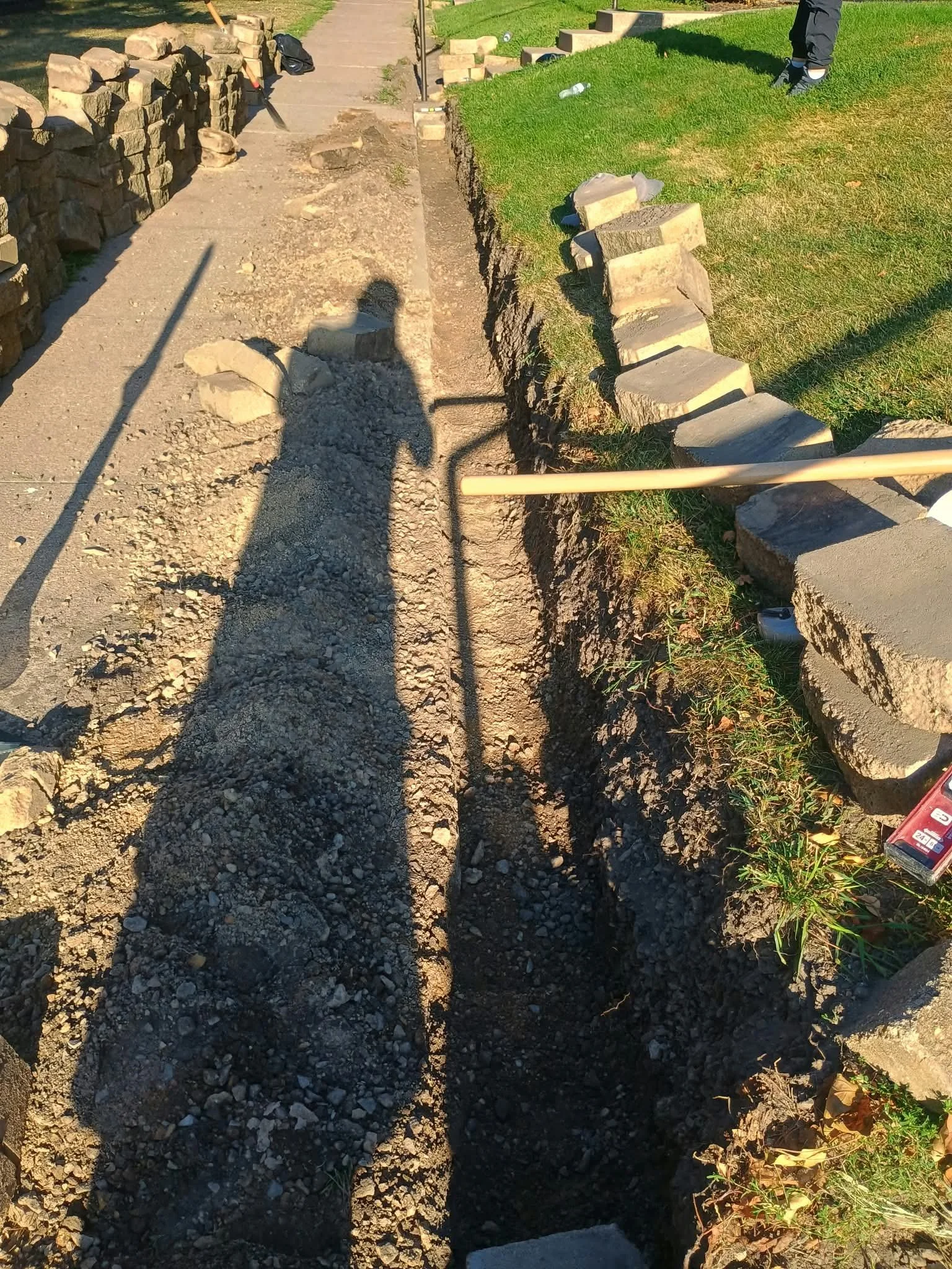 A sidewalk under construction with a trench dug along the side, rocks and dirt scattered in the trench, and stone blocks on the left. There is a grassy area on the right with some bricks and construction tools.