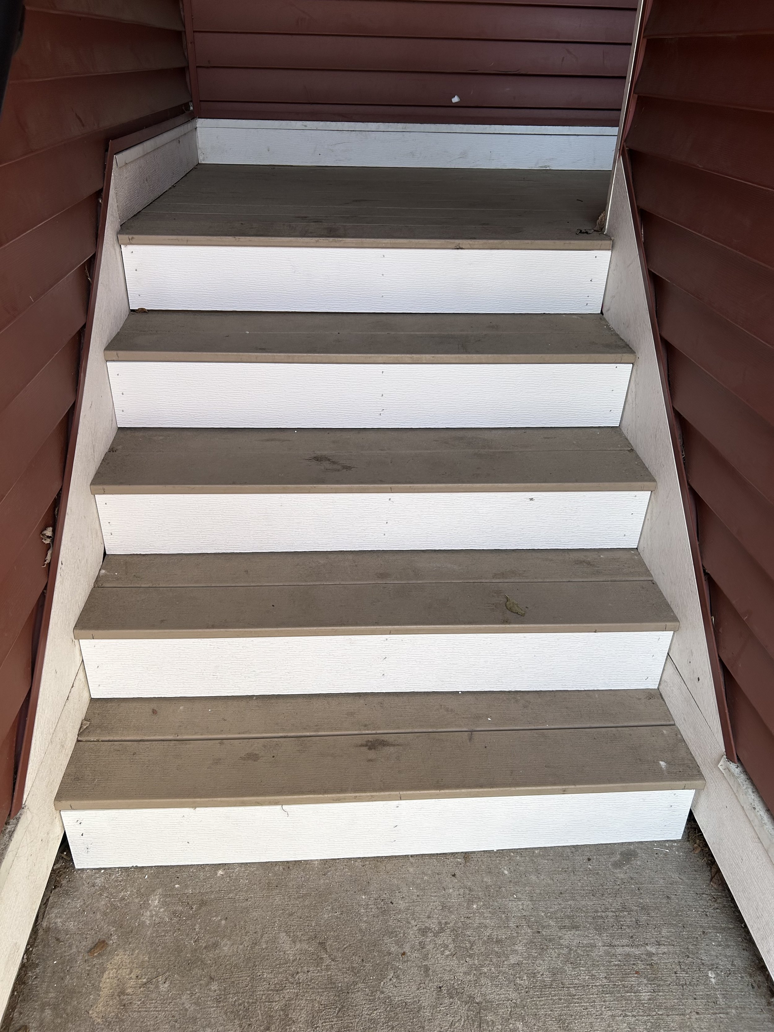 A set of four outdoor stairs with brown painted treads, white risers, and a concrete porch at the top, flanked by reddish-brown wooden siding.