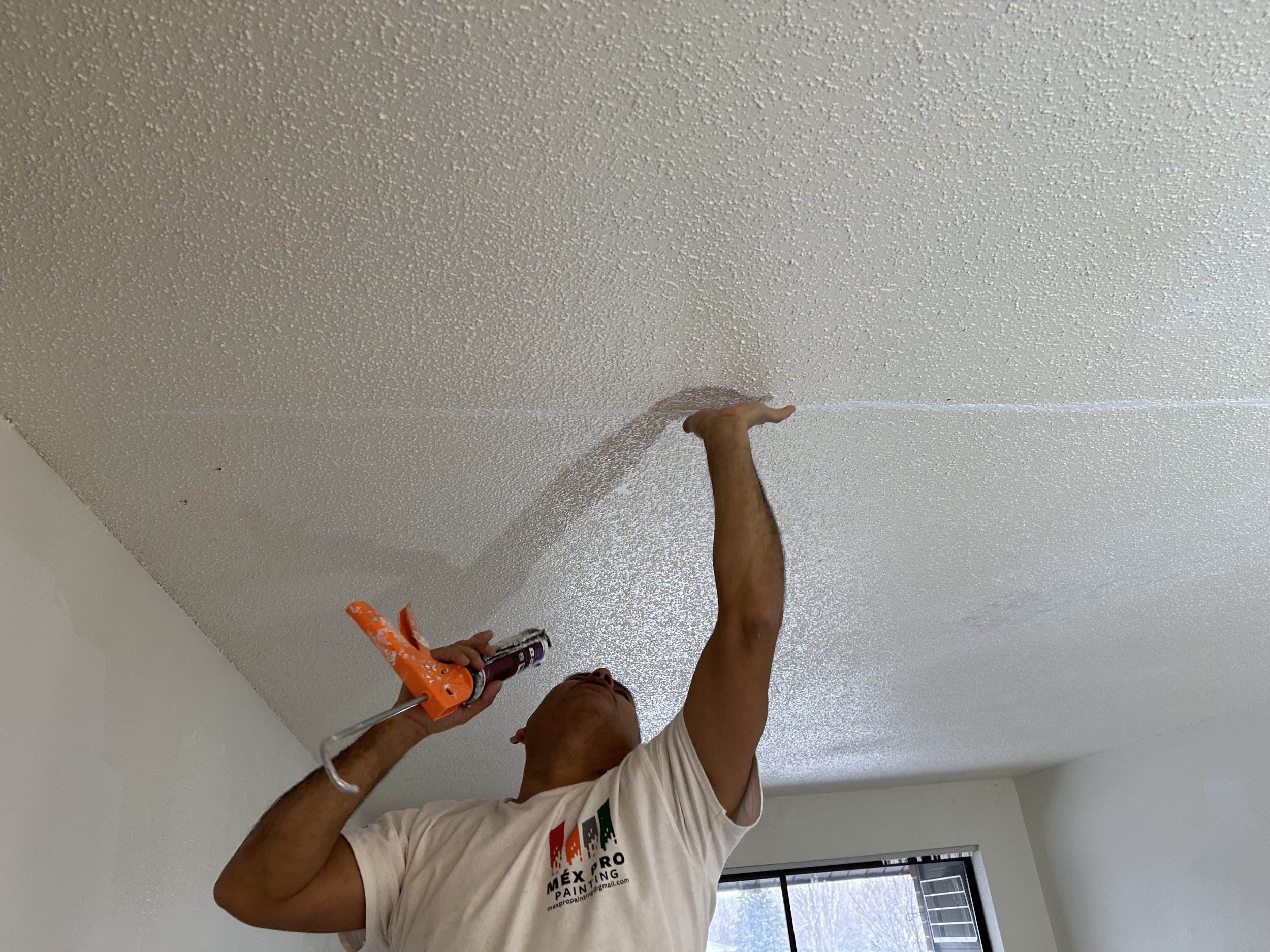Man applying textured ceiling paint with a sprayer in a room with white walls and window.