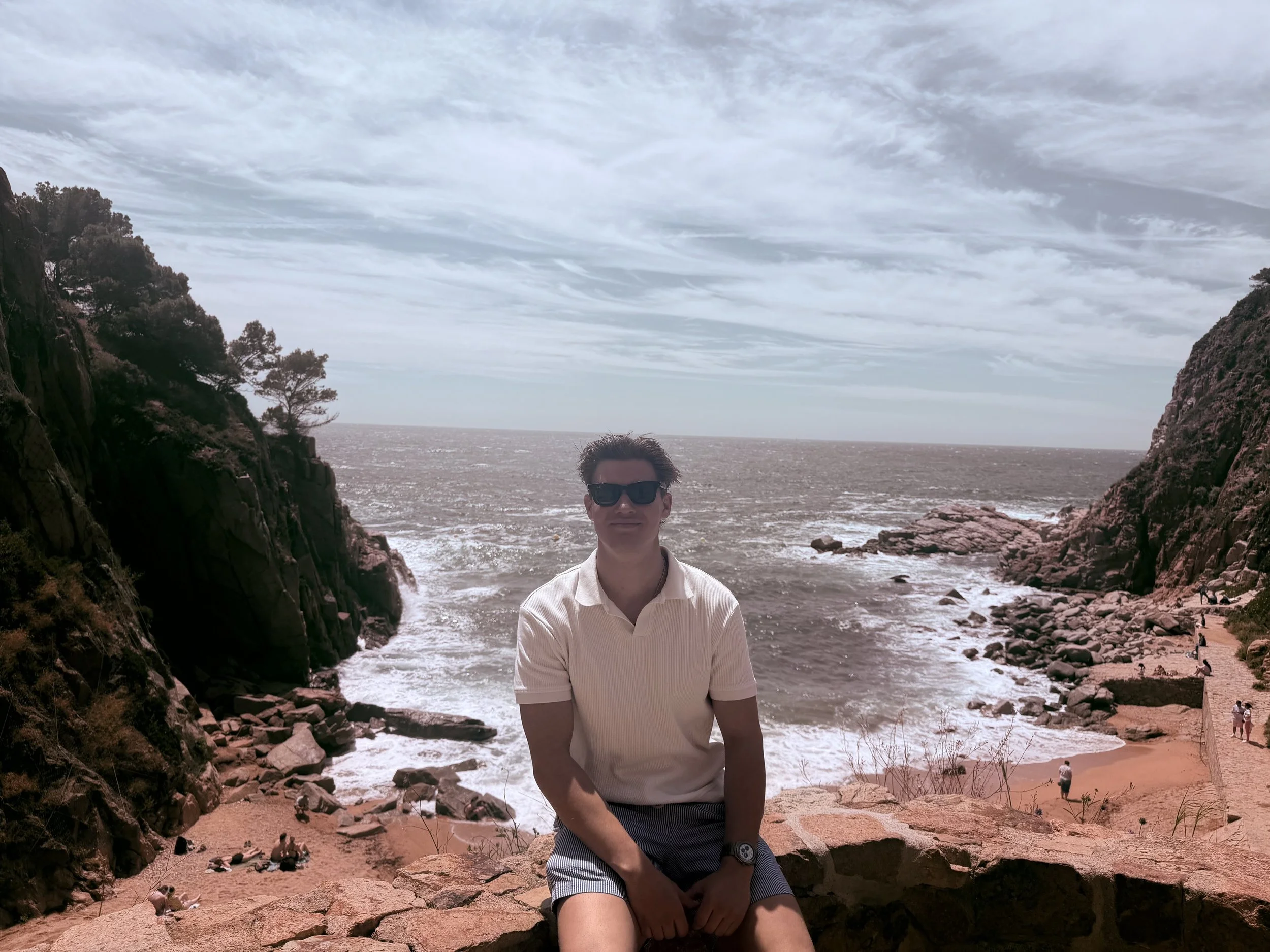 A man wearing sunglasses and a white shirt sitting on rocks at a beach with cliffs on either side and the ocean in the background.