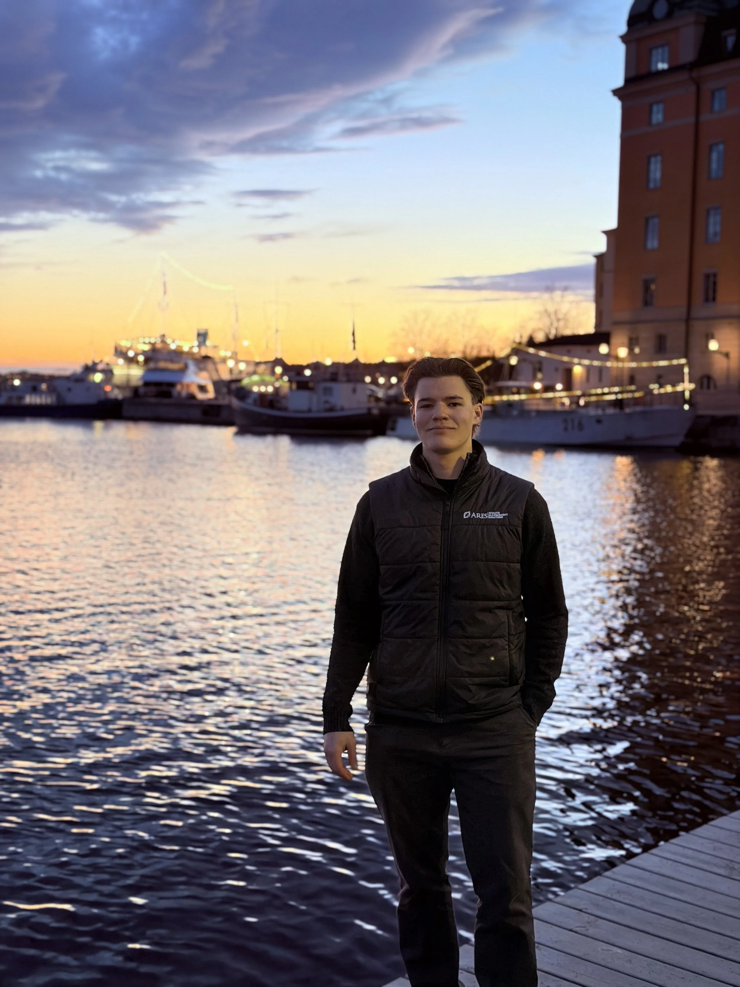 A young man standing on a wooden dock by the water during sunset with boats, a building, and a sky with clouds in the background.