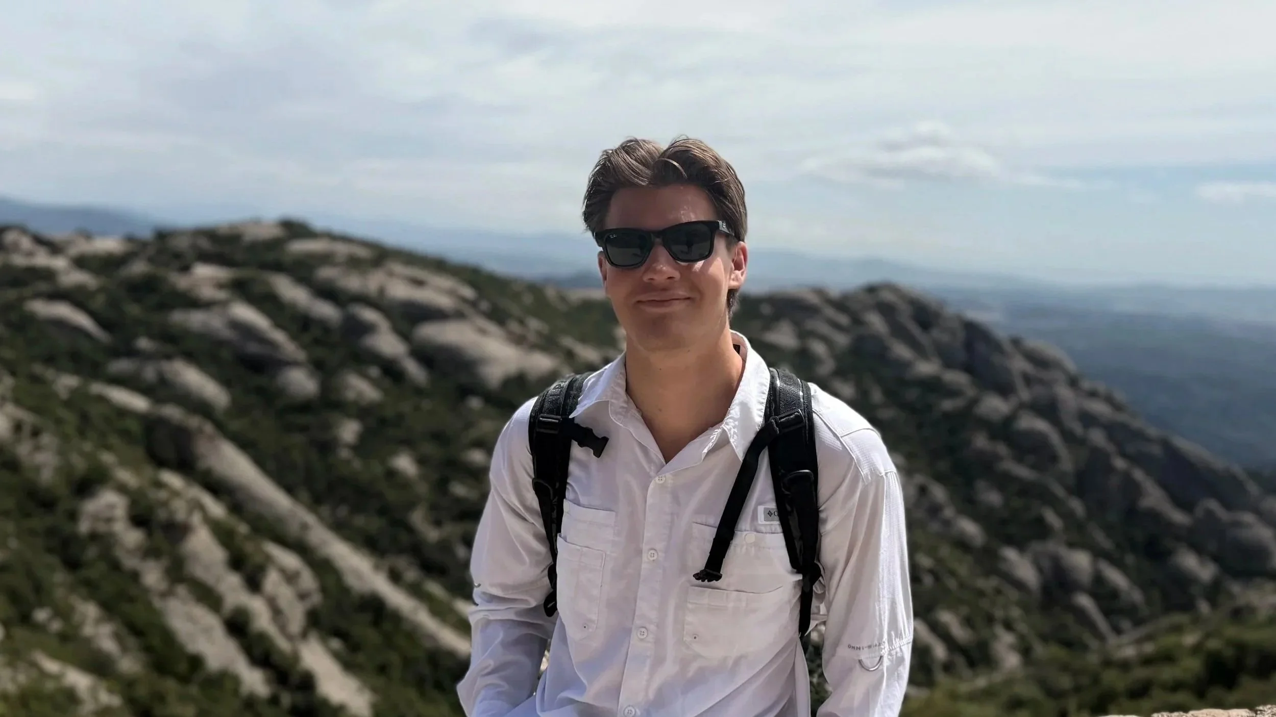 A young man with brown hair wearing sunglasses, a white shirt, and a backpack, standing outdoors on rocky terrain with a mountain landscape in the background.