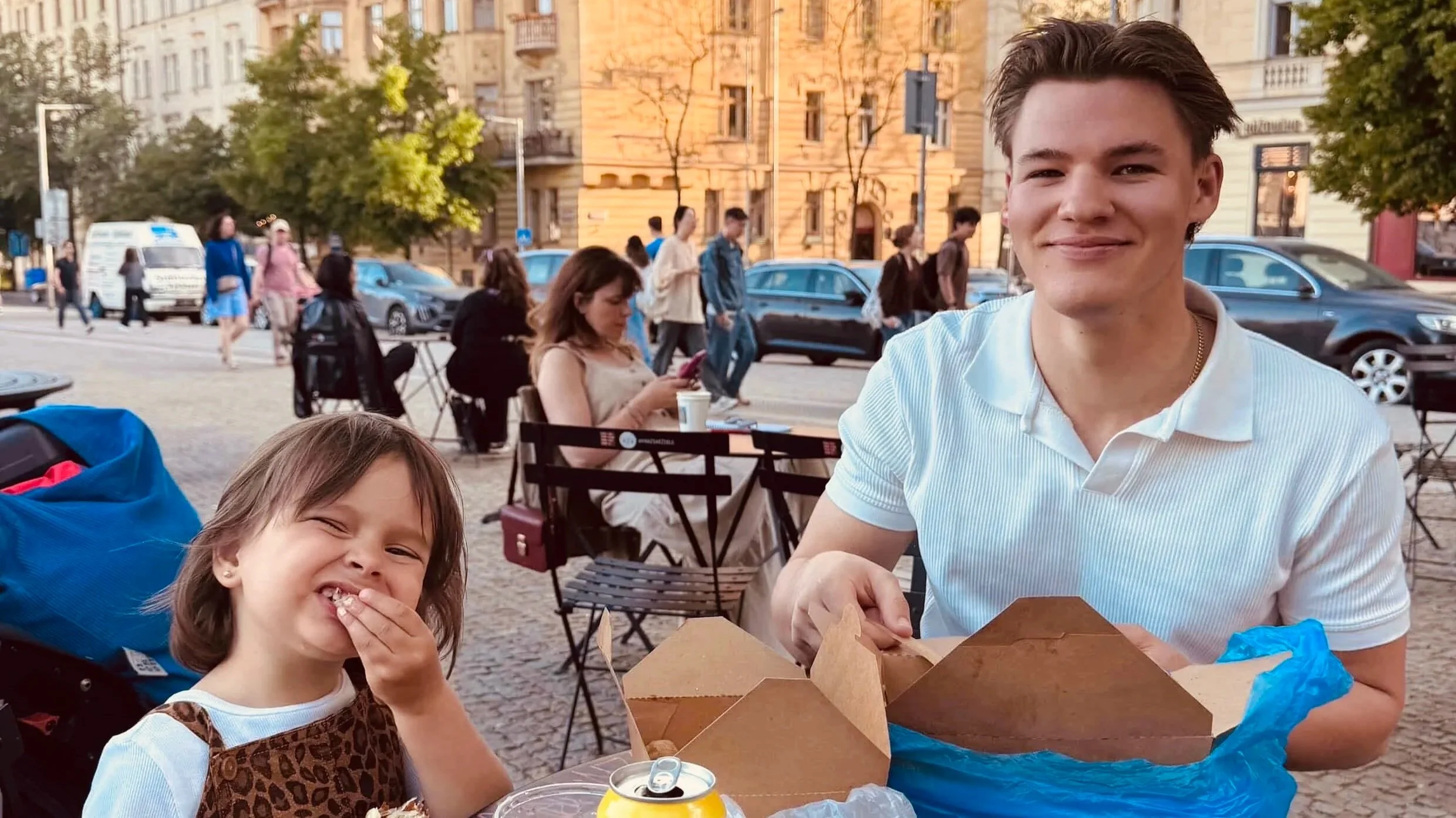 A young girl with brown hair smiling and eating, sitting outdoors at a table with a man in a white shirt. The background shows people walking on the street, trees, and buildings.