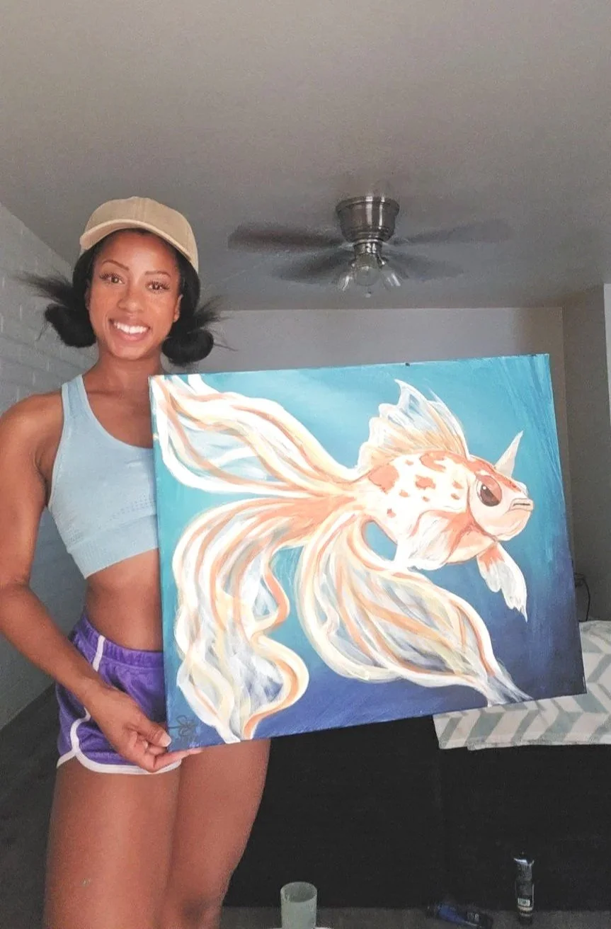 Young woman with a beige cap and purple athletic shorts holding a painting of a sea creature with flowing pink and orange fins, set indoors.