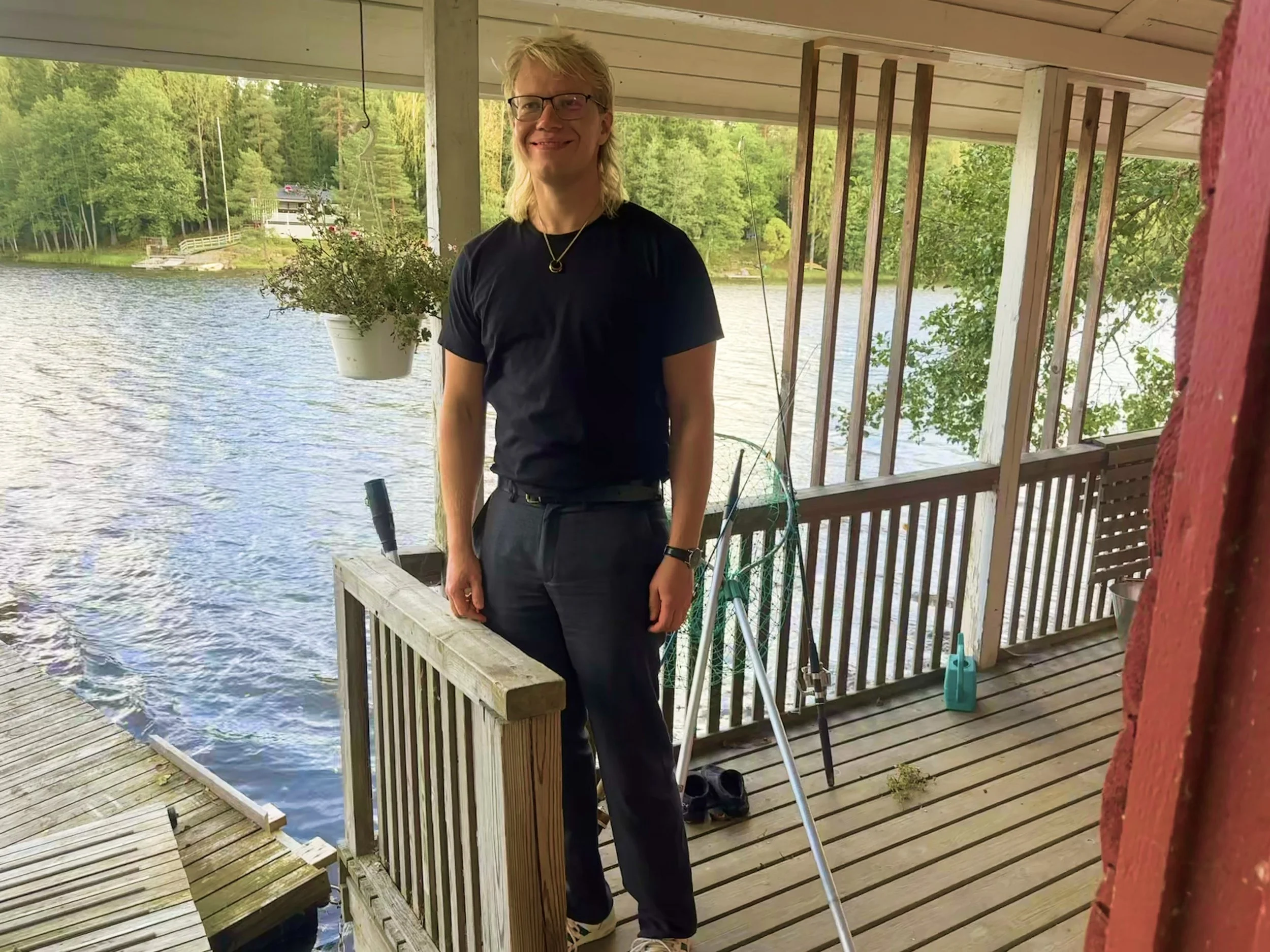 A man with glasses and long blond hair smiling and standing on a wooden dock by a lake, with trees in the background, and fishing gear beside him.