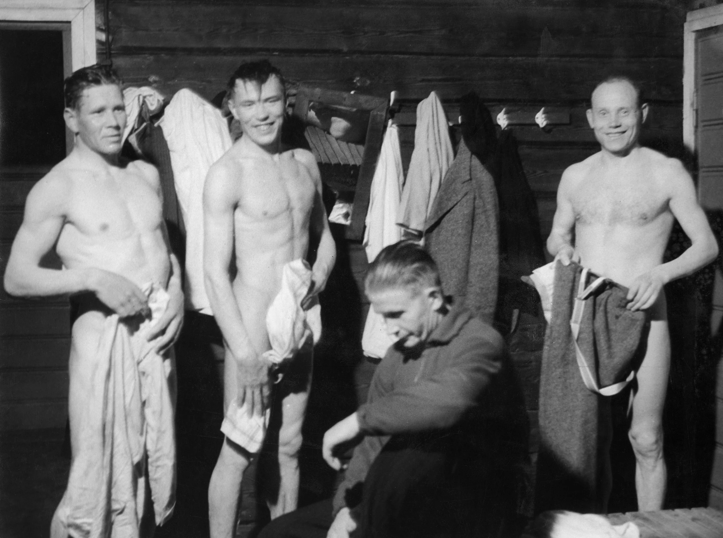 Black and white photo of three shirtless men standing indoors in what appears to be a wooden cabin or locker room. Two men are smiling at the camera, while the third man in the foreground appears to be looking down. Clothes are hanging on hooks behind them.