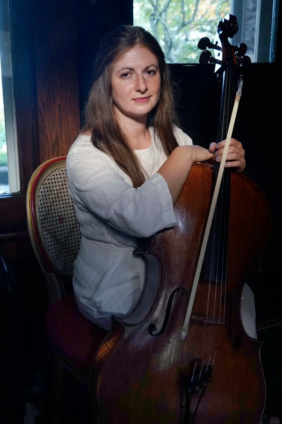 A woman with long brown hair playing the cello indoors by a window.
