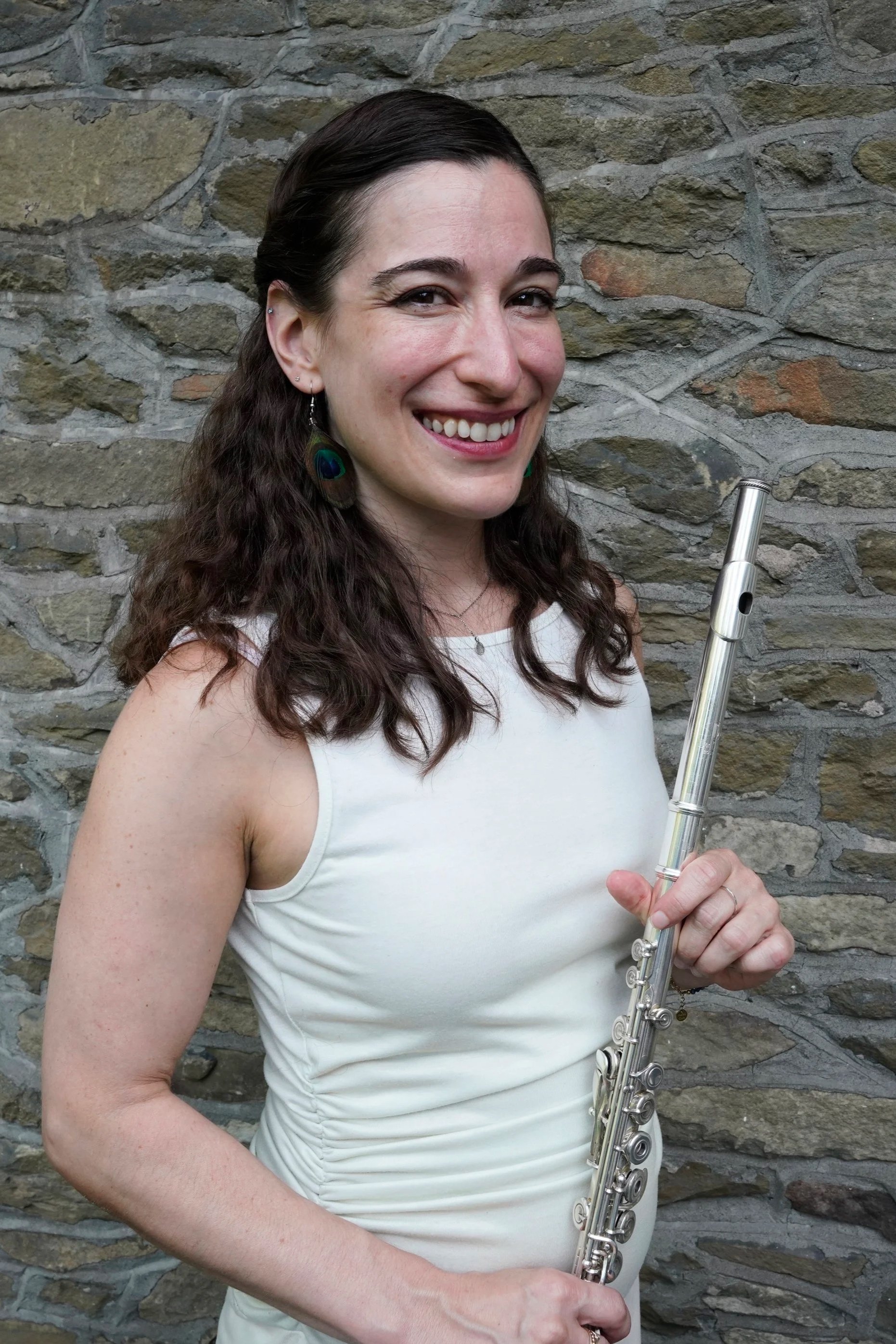 A woman with long curly brown hair, wearing a white sleeveless top, smiling and holding a silver flute, standing in front of a stone wall.