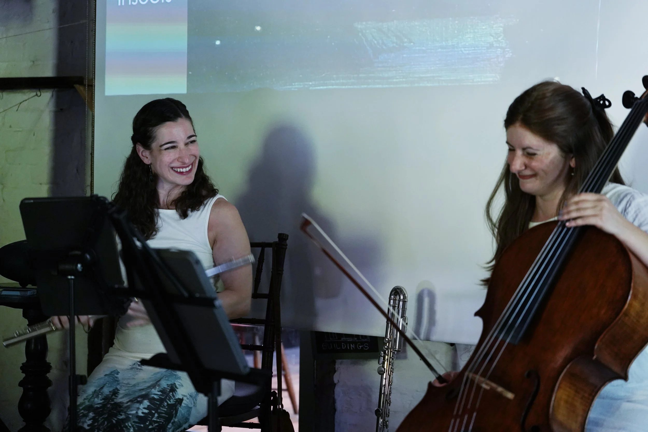 Two women sitting and smiling, one playing a cello, in a room with a white wall and a projected screen behind them.
