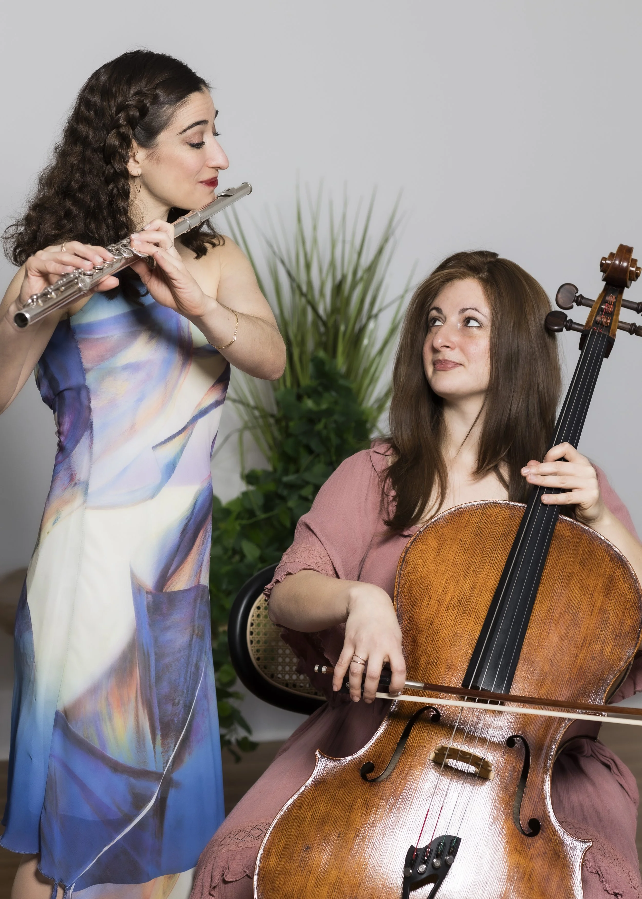 Two women, one playing the cello and the other playing the flute, are facing each other indoors with a plant in the background.