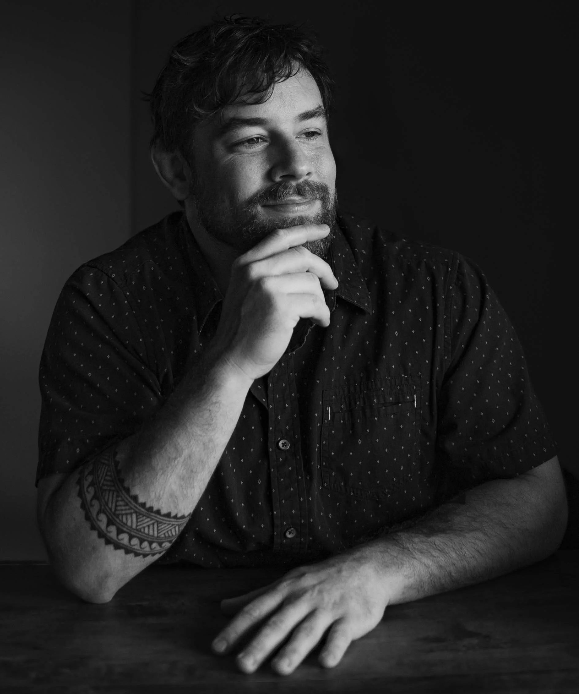 Black and white photo of a man with a beard and short wavy hair, resting his chin on his hand and looking thoughtfully to the side.