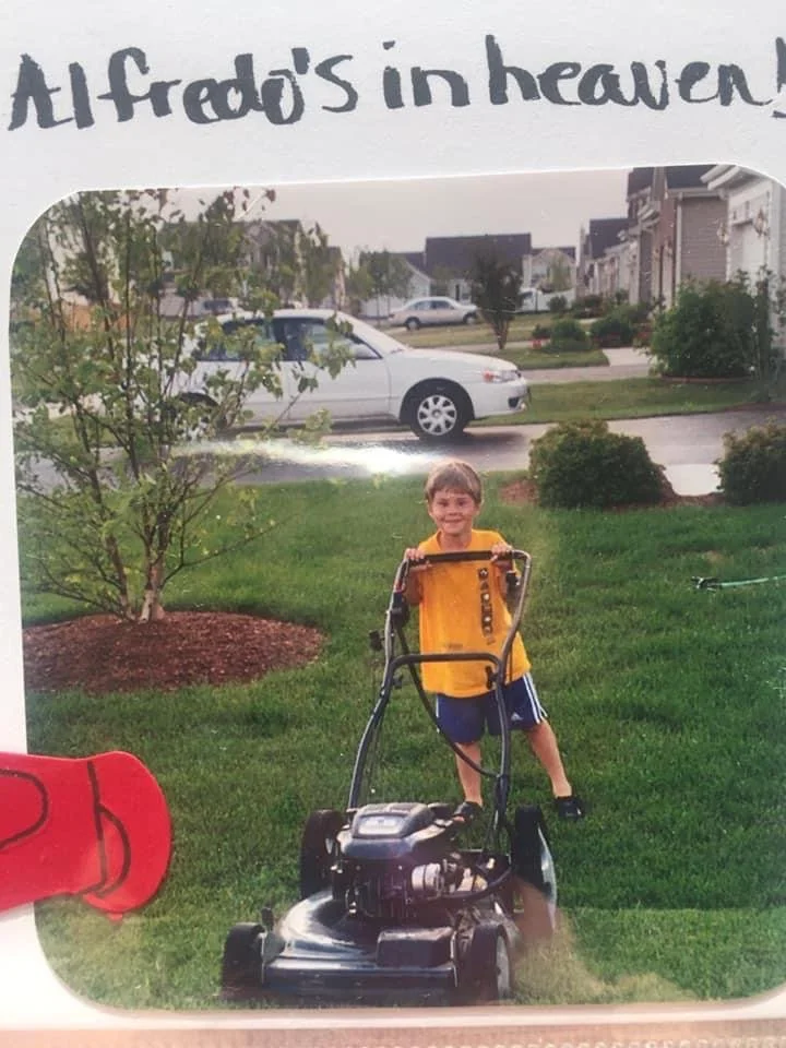 A young boy in a yellow shirt and blue shorts mowing the lawn in a suburban neighborhood.