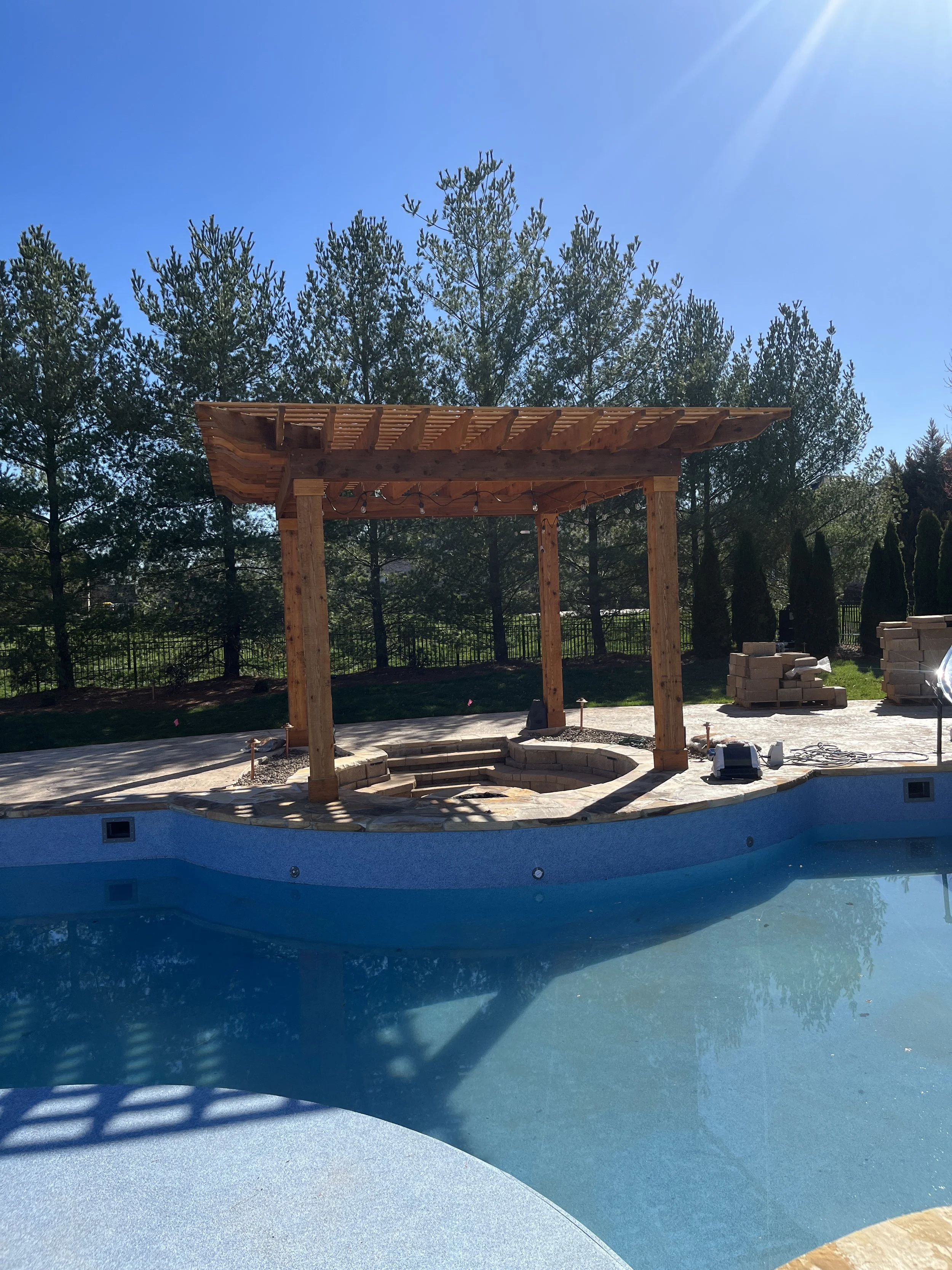 A backyard scene with a pool and a wooden pergola. The pool has a curved edge, and there are stacks of bricks and construction tools nearby, indicating ongoing construction or renovation. Tall trees and a clear blue sky are in the background.