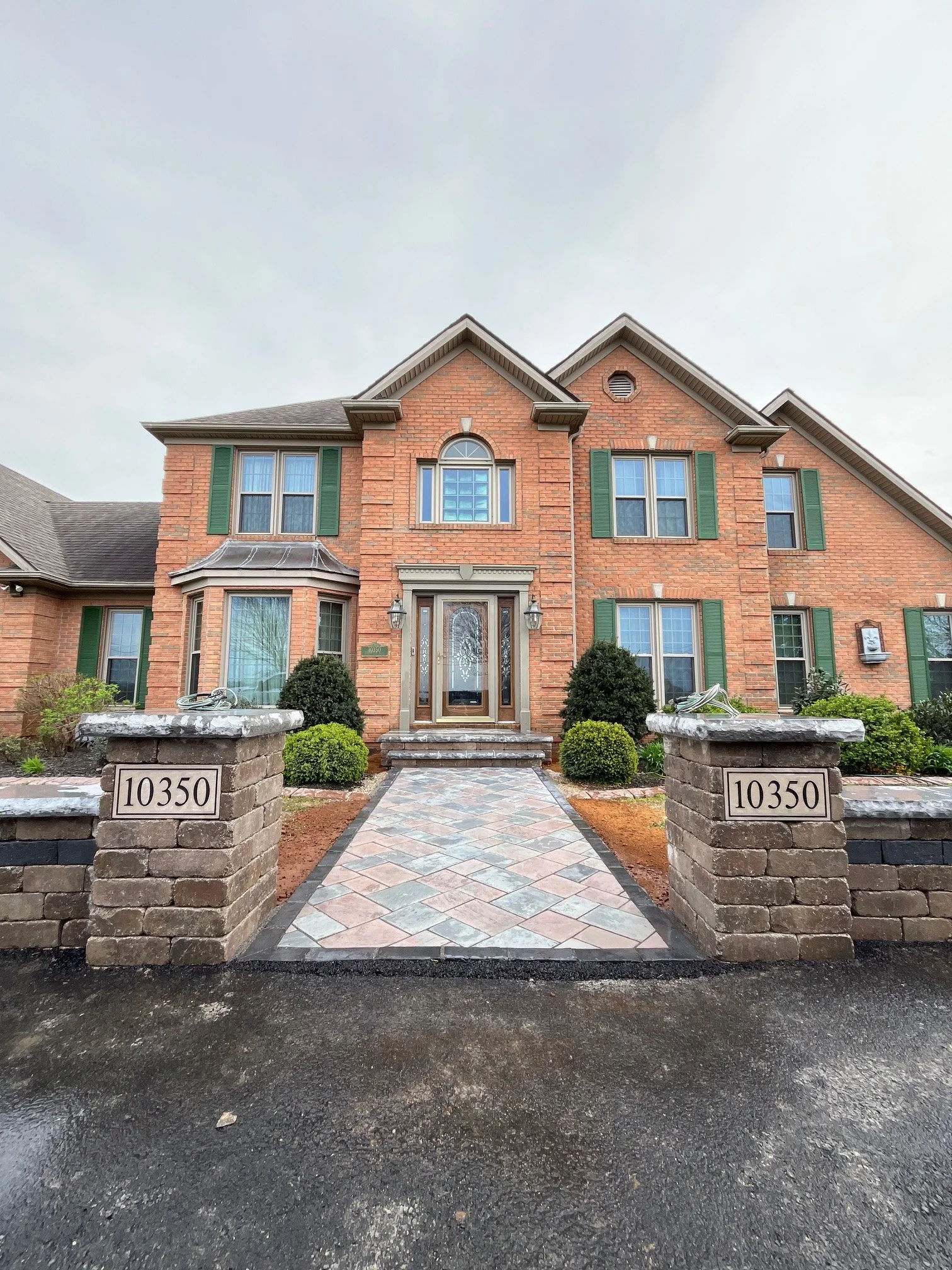 Front view of a large brick house with green shutters, multiple windows, and a decorative front door. The house has a pitched roof and a small front yard with well-kept bushes. Two brick pillars with house numbers 10350.5 and 10350.0 mark the entrance, and a paved walkway leads to the front door. The ground in front appears wet and dark, possibly from recent rain.