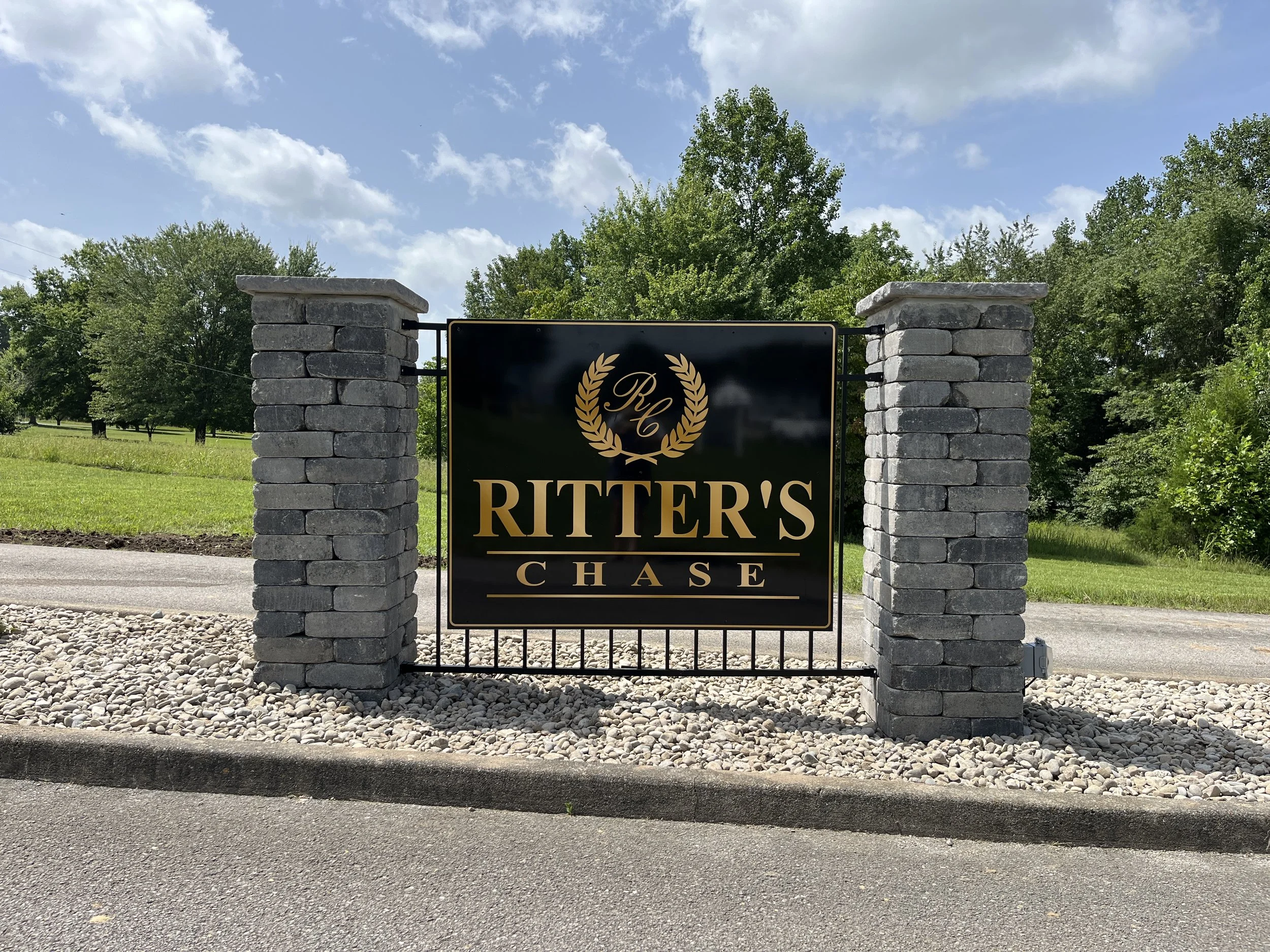 A black sign with gold lettering and a laurel wreath, reading 'Ritter's Chase,' is mounted between two stone pillars on a gravel base, with trees and blue sky in the background.