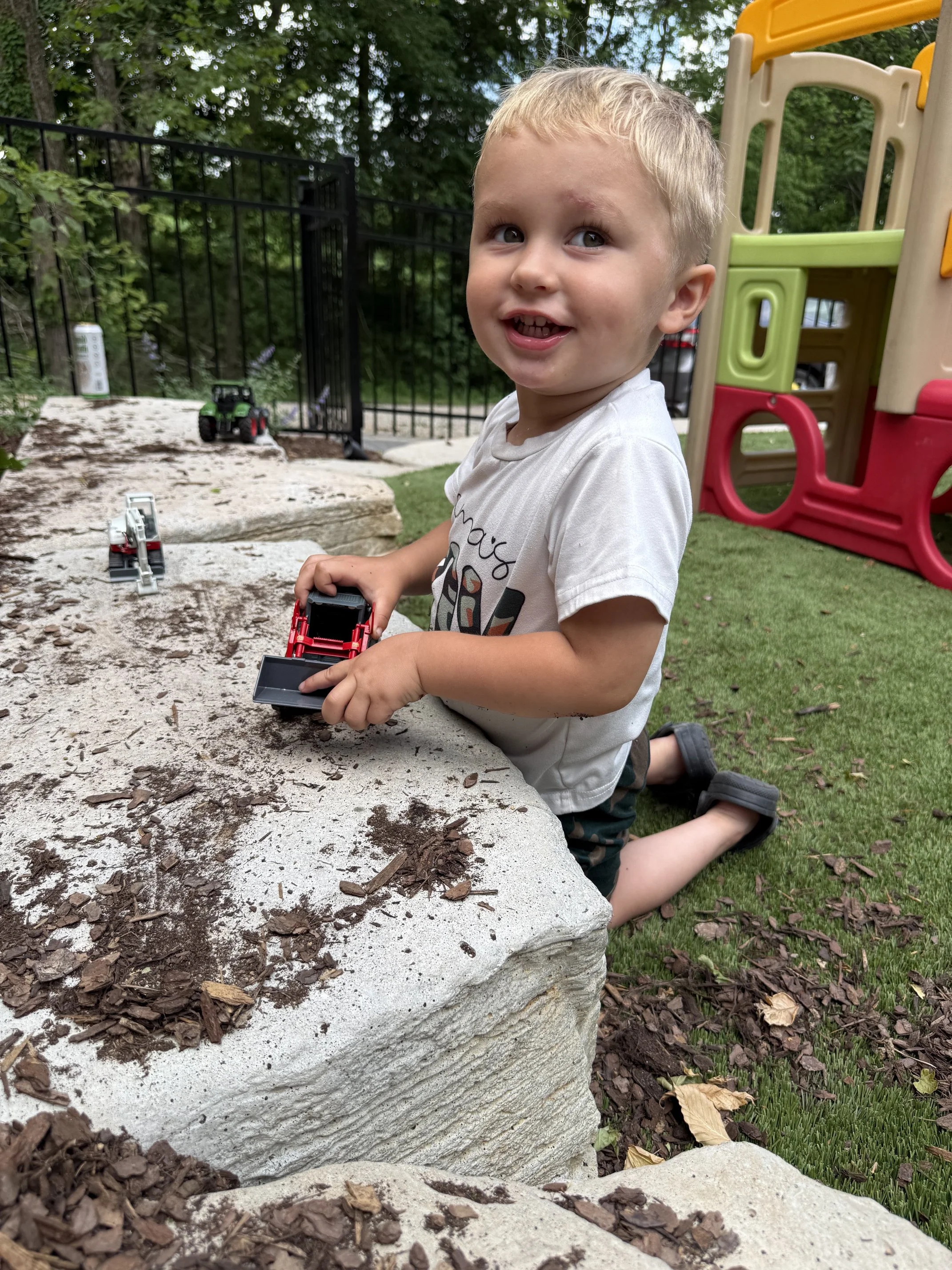 A young boy with blonde hair kneeling on the grass, playing with toy vehicles near a stone wall in a backyard, with trees and playground equipment in the background.