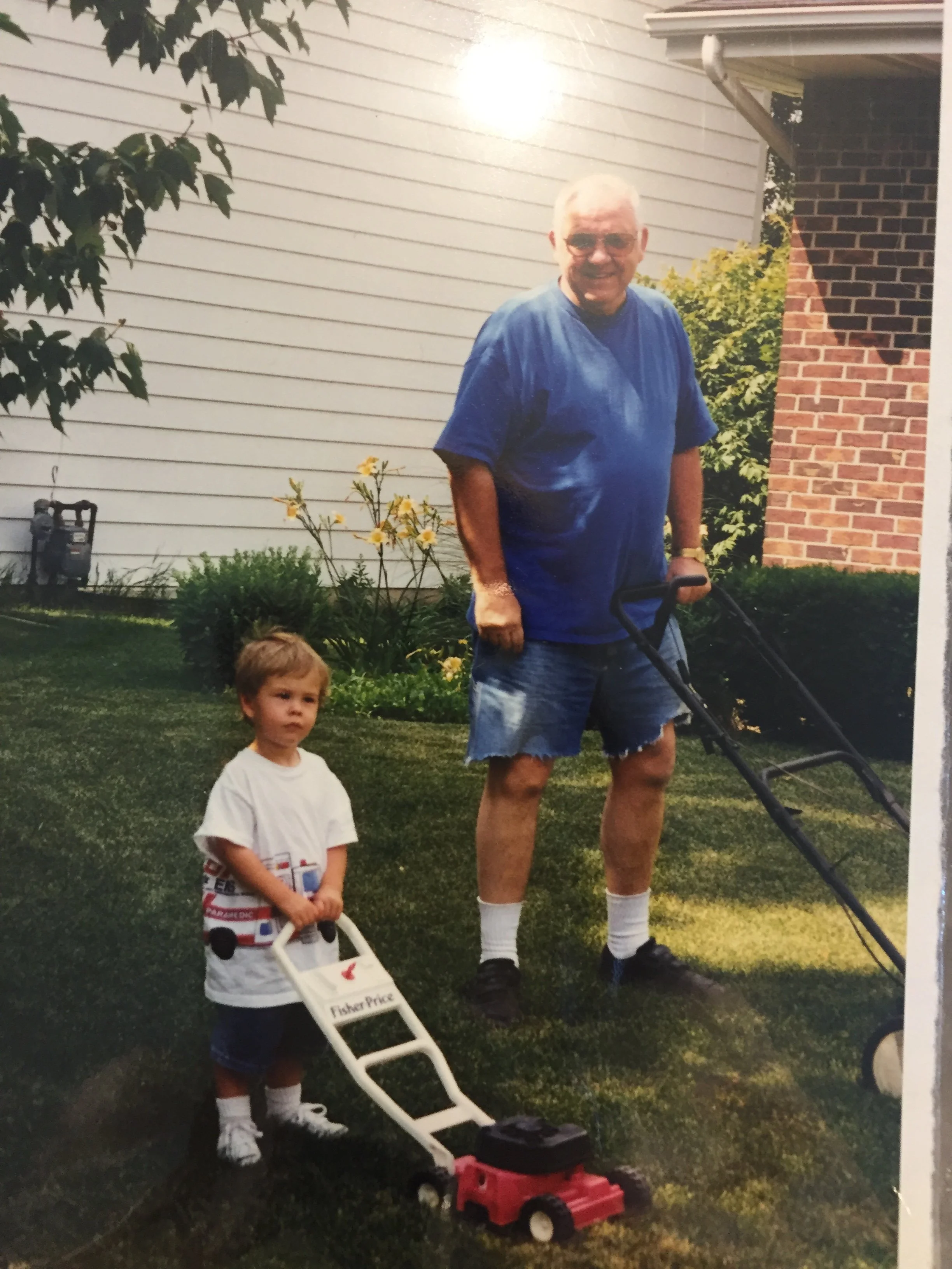 A grandfather in a blue shirt and shorts mowing the lawn with a push mower while a young boy stands nearby holding a toy lawn mower. The scene is in a backyard with grass, flowers, and a house with white siding and brick wall.