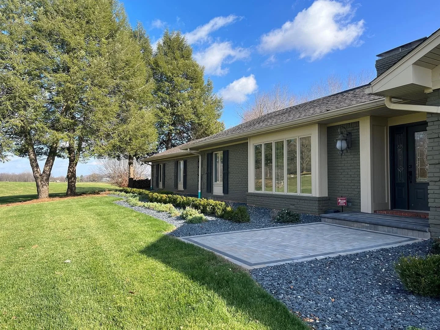 Front yard of a house with a patio, green lawn, trees, and a bright blue sky.