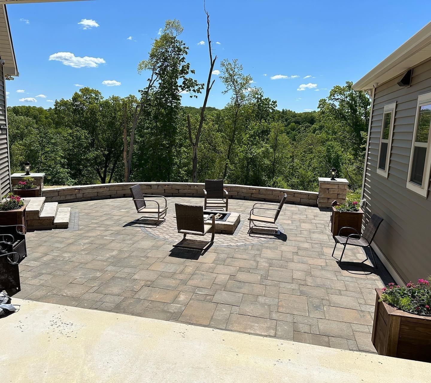 A spacious backyard patio with a stone stone surface, outdoor chairs arranged around a fire pit, and potted plants along the edge. In the background, there are trees and a clear blue sky with a few clouds.