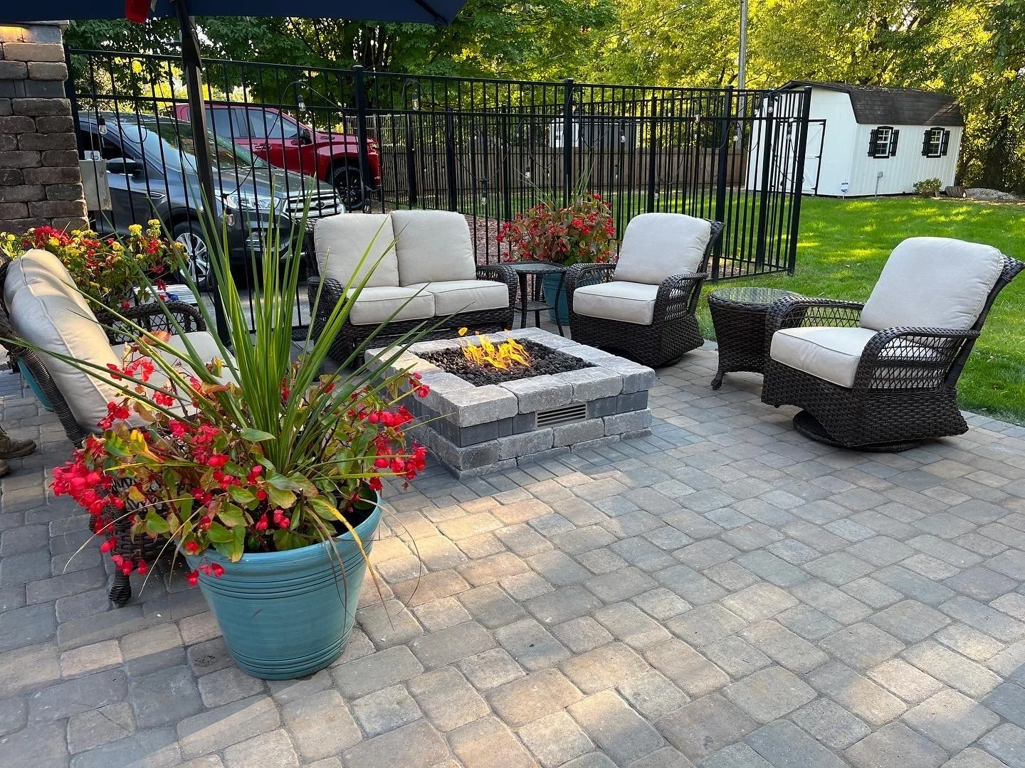 Outdoor patio with wicker furniture, including three armchairs and a loveseat with cream cushions, surrounding a firepit made of bricks. There are potted plants with red flowers on the patio. In the background, there is a fenced yard, a small white s