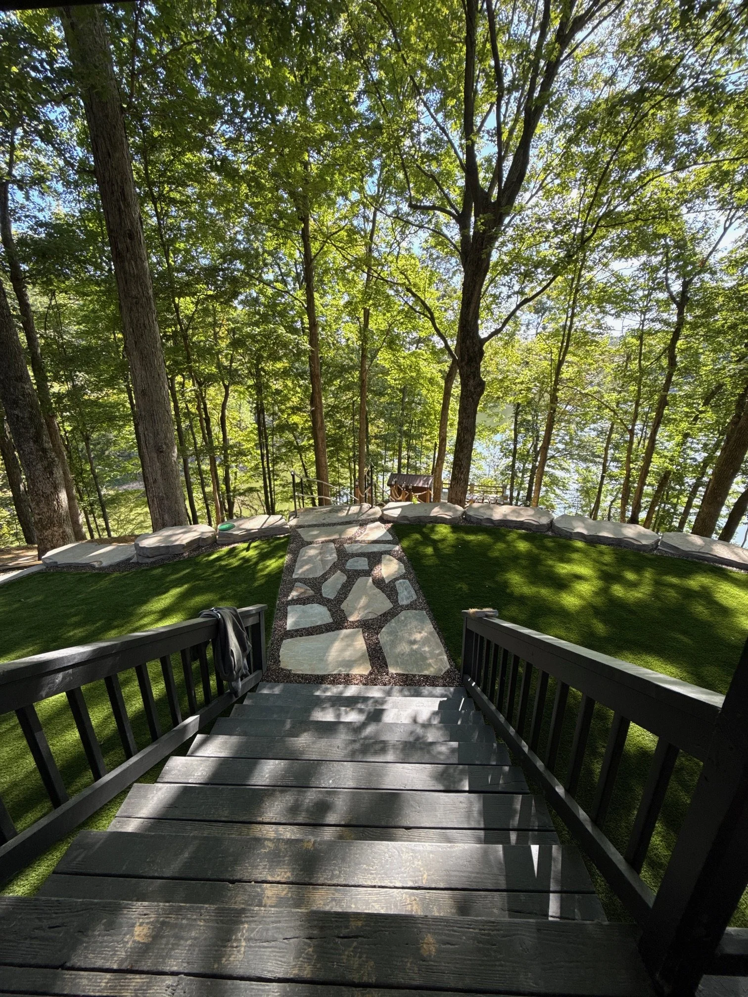 A wooden staircase leading down to a grassy area in a forest, surrounded by tall trees with green leaves and a small wooden structure in the distance.
