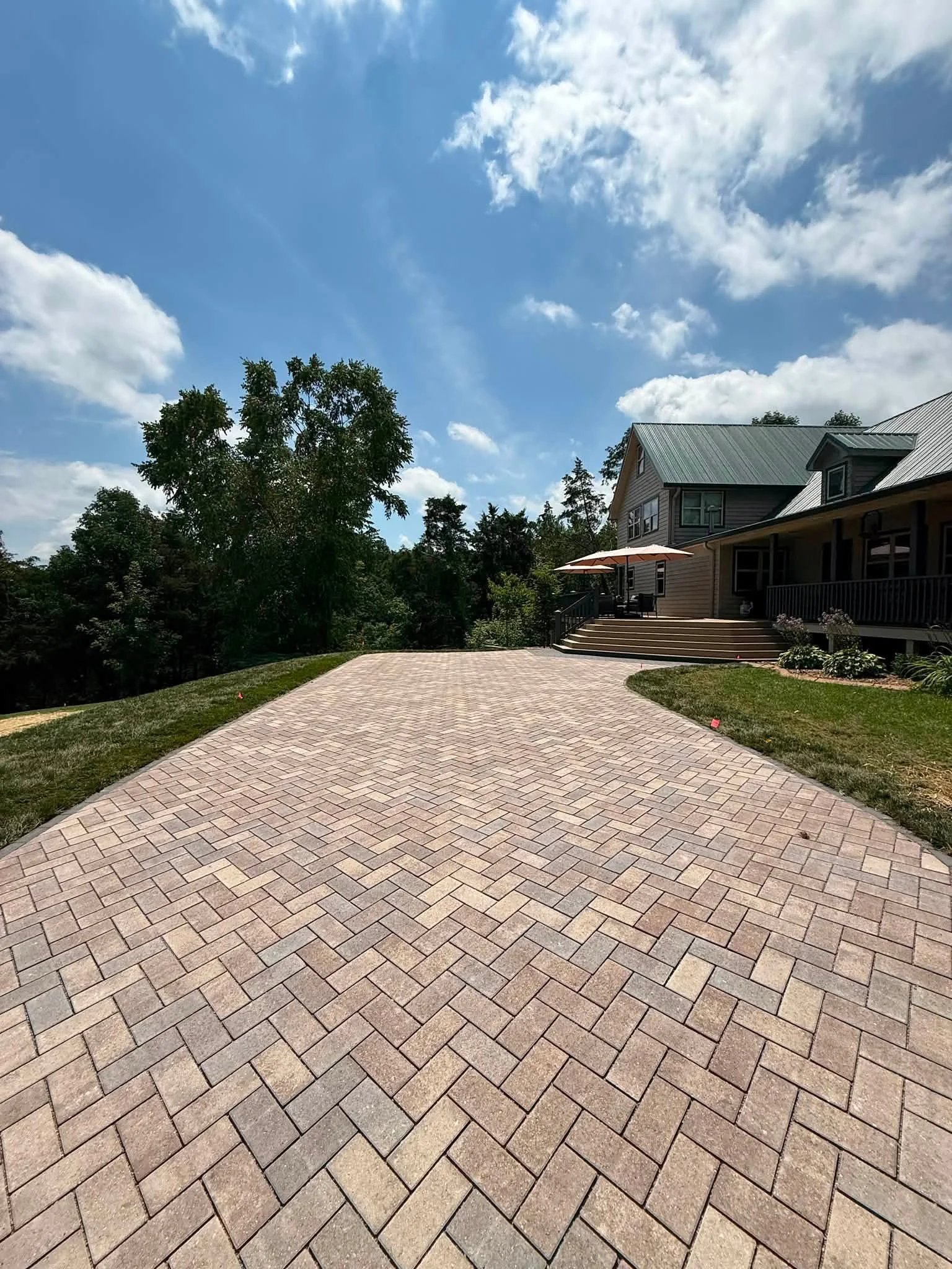 Paved brick driveway leading up to a house with a porch and umbrella, surrounded by trees and a blue sky with clouds.