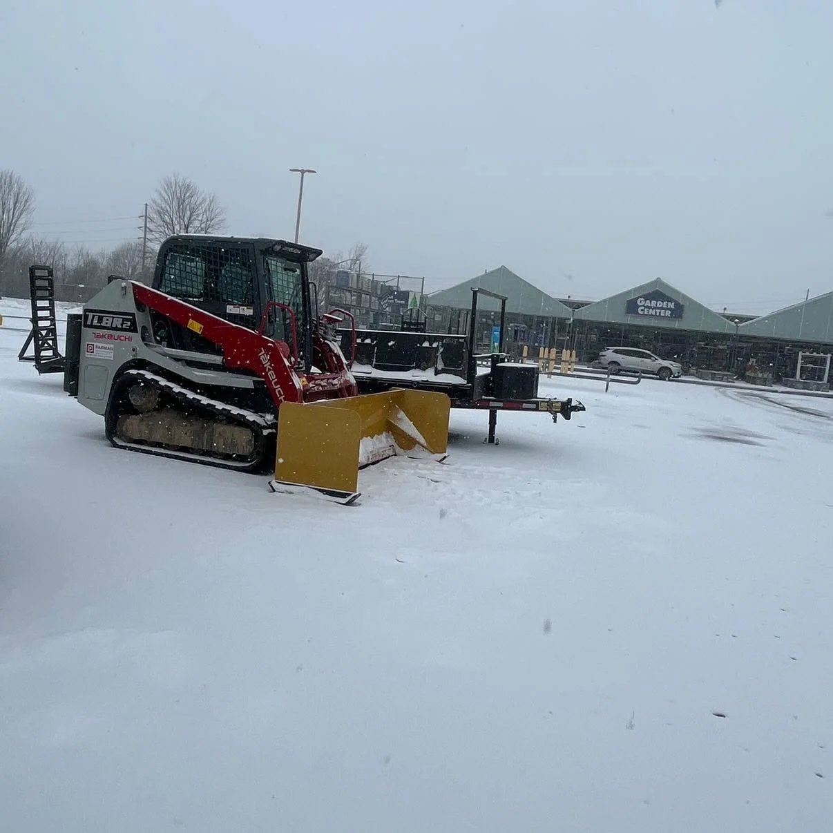 Snow-covered parking lot with a skid-steer loader and a wheelbarrow near a garden center store.