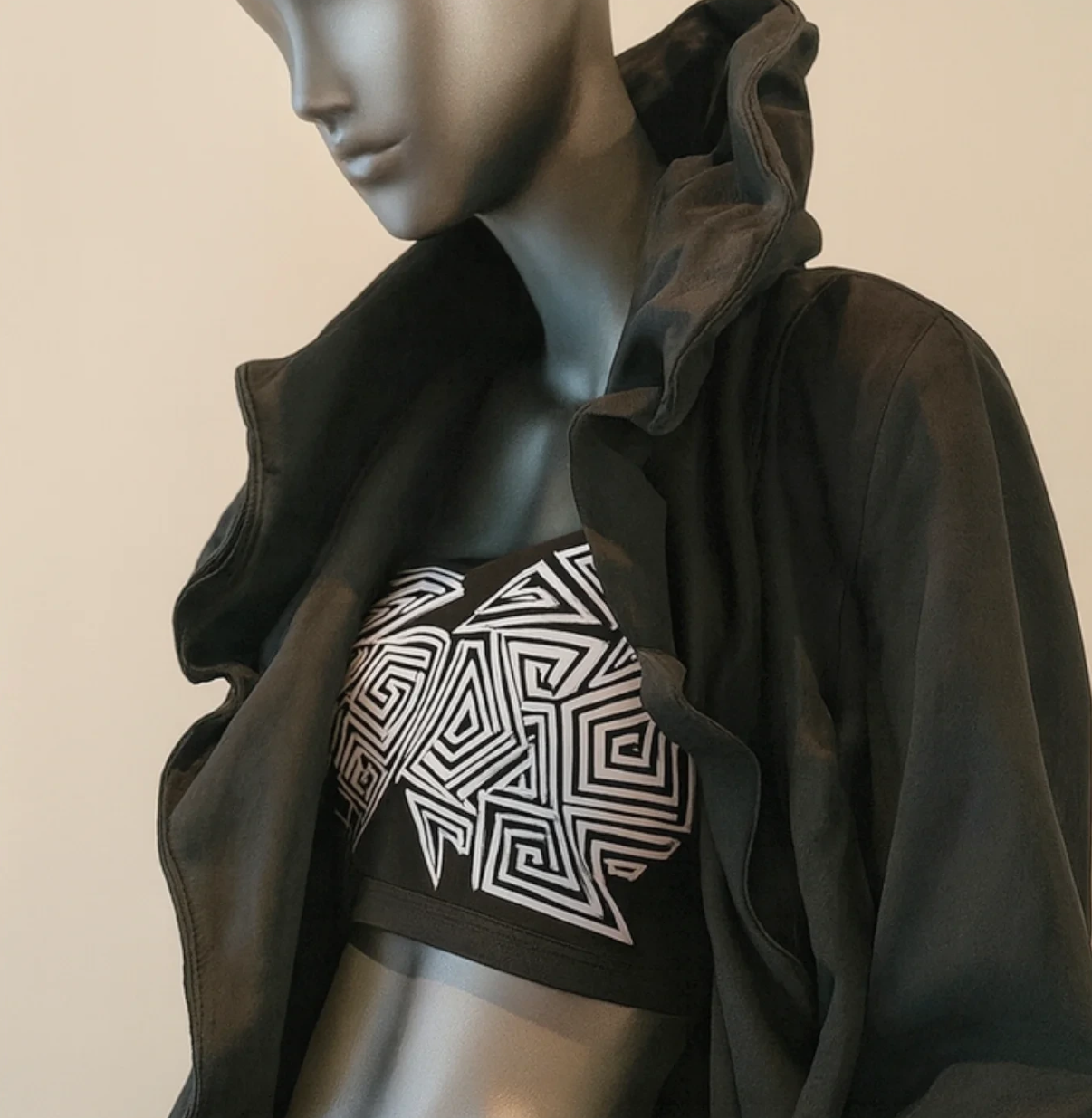 A mannequin dressed in a black hoodie with the hood up, layered over a black and white geometric patterned crop top.