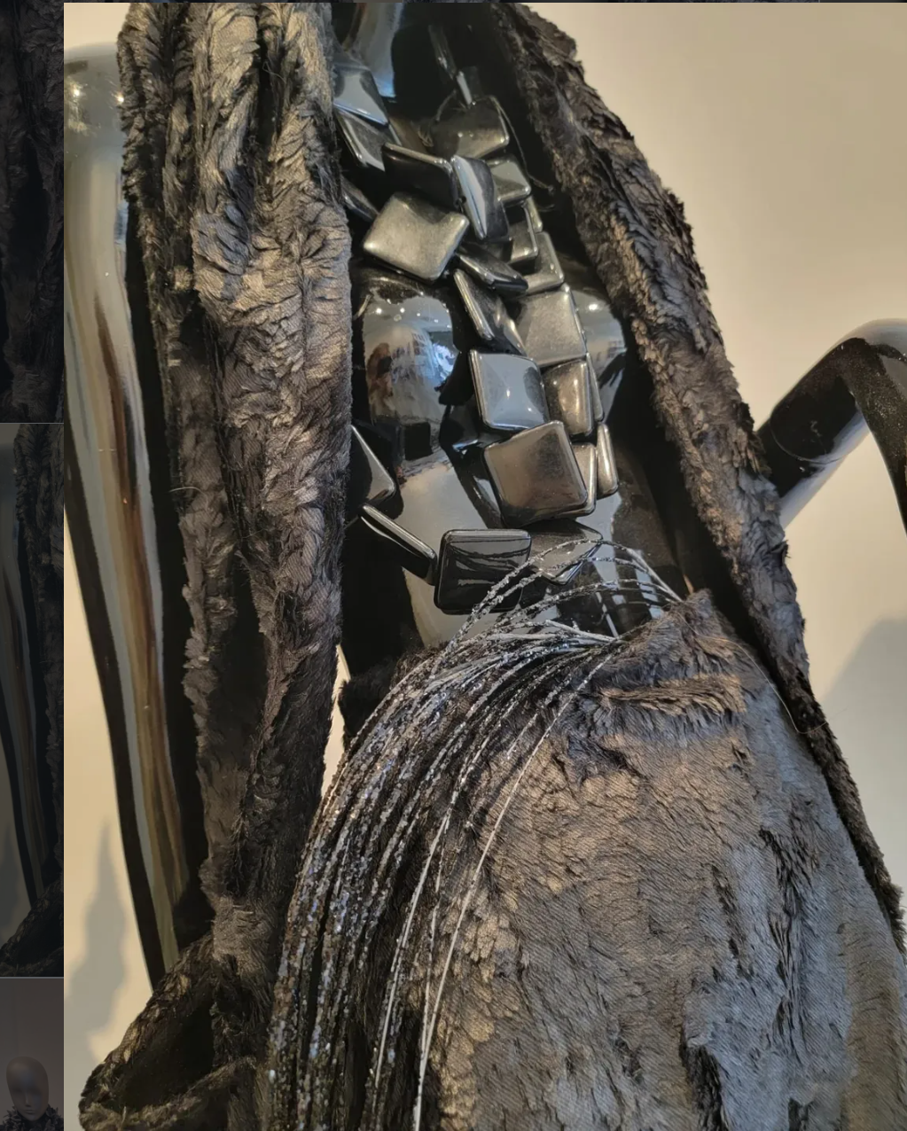 Close-up of a mannequin with a dark, textured fur coat, a shiny black leather outfit, and a necklace of large, square, reflective black beads.